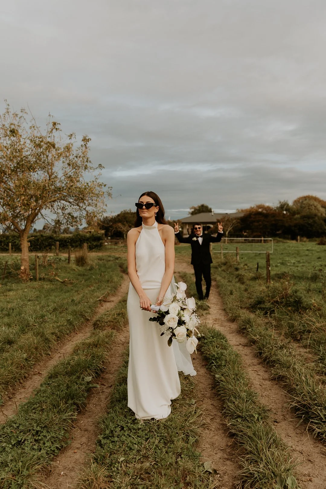 A woman in a white wedding dress holding a bouquet of white flowers stands on a dirt path in a rural field, with a man in a black tuxedo in the background making peace signs.