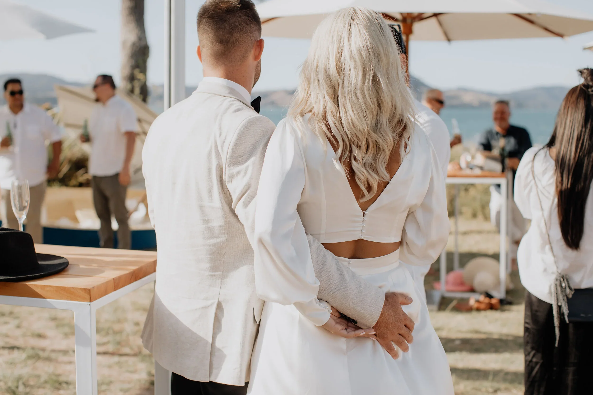 A couple, dressed in wedding attire, stands close together at an outdoor beach wedding ceremony, with guests and water in the background.