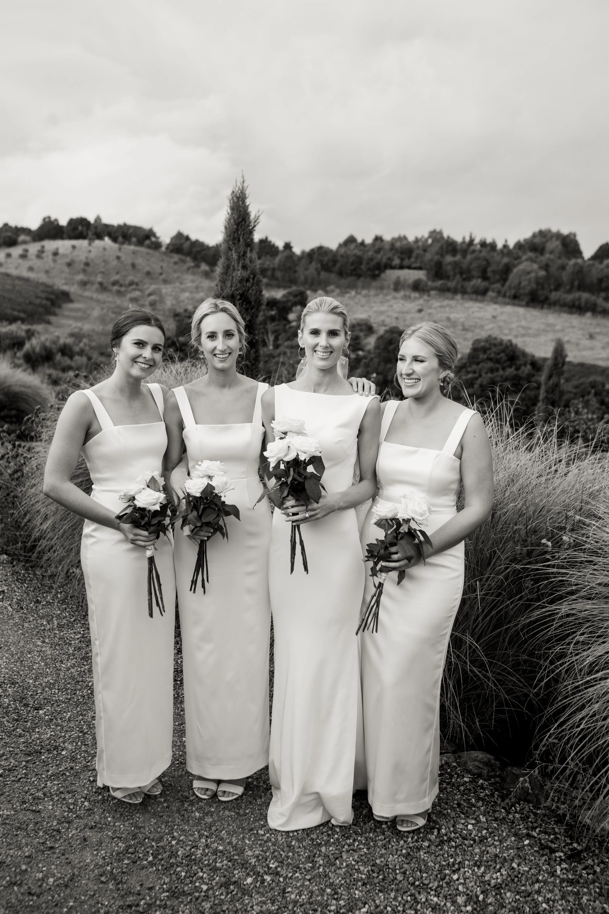 Four women in white dresses holding bouquets of flowers, standing outdoors on a gravel path with rolling hills and trees in the background.