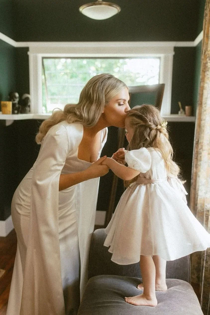 A woman in a white dress kisses a young girl in a white dress on the forehead, inside a room with a window and natural light.