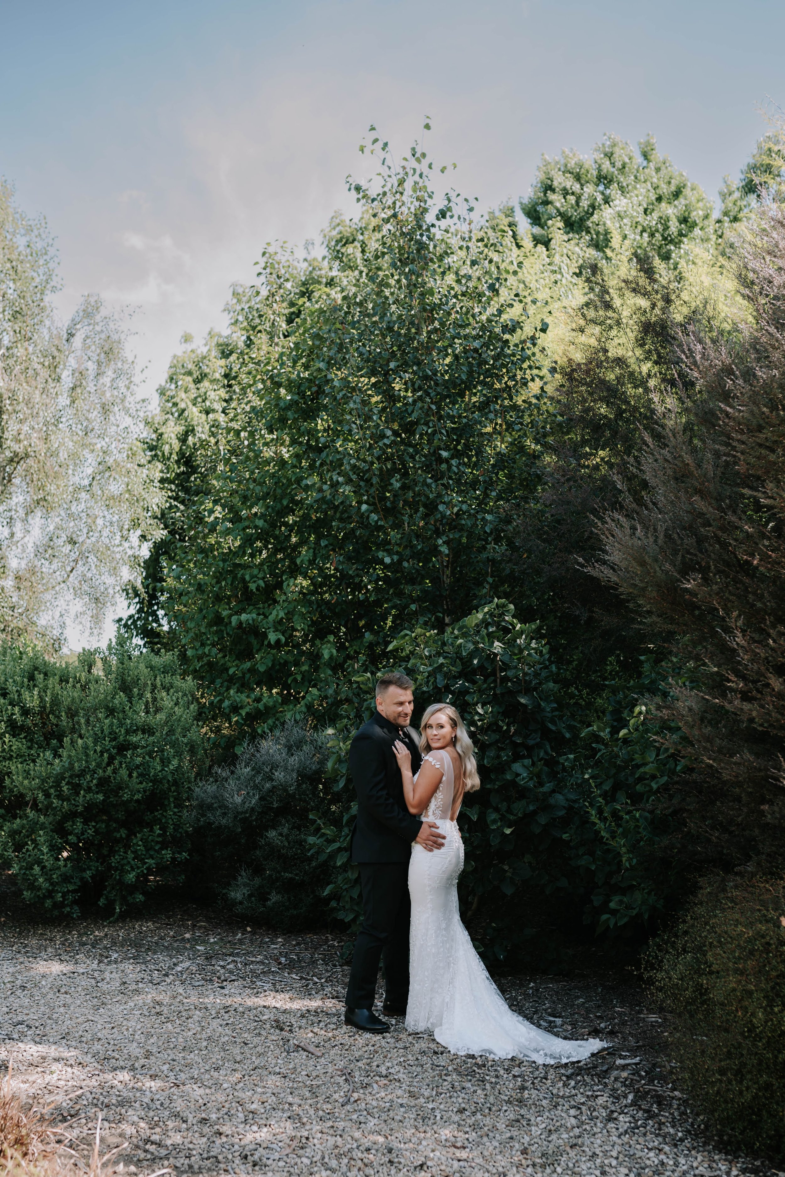 A bride and groom standing outdoors on a gravel path surrounded by lush green trees, with the bride in a white wedding dress and the groom in a black suit.
