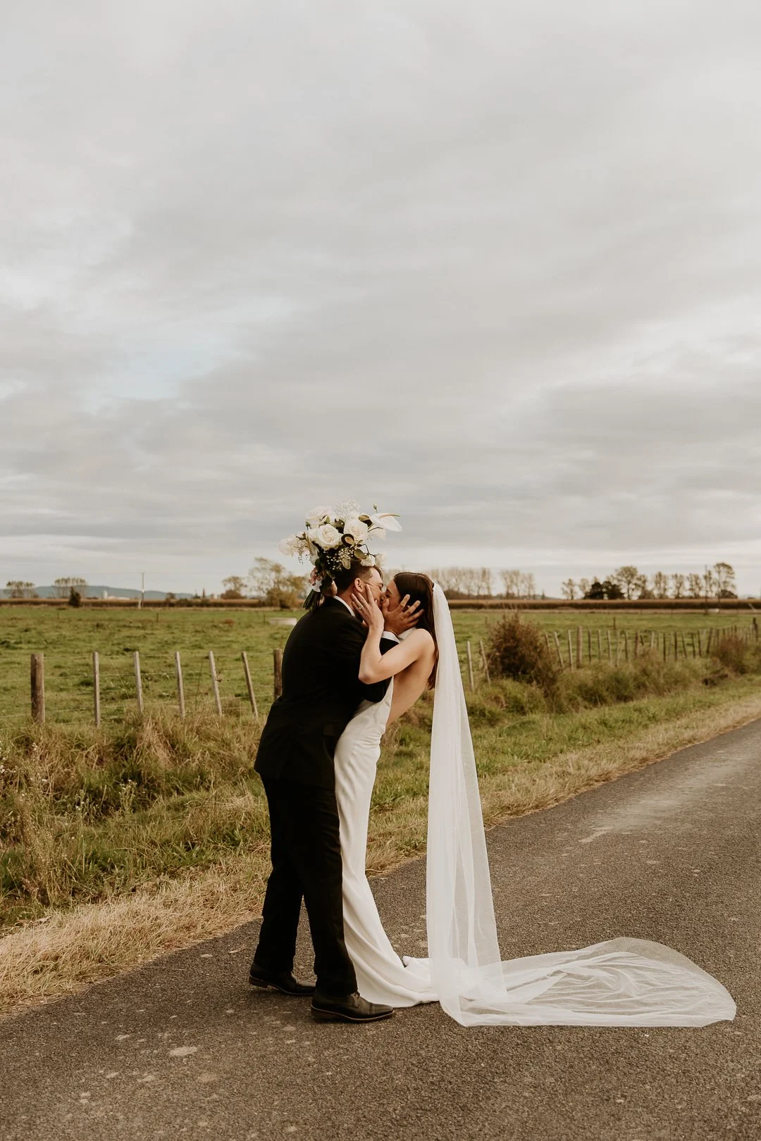 A couple in wedding attire sharing a kiss and embrace on a rural road, with a large bouquet of flowers on the groom's head, fields, and a cloudy sky in the background.