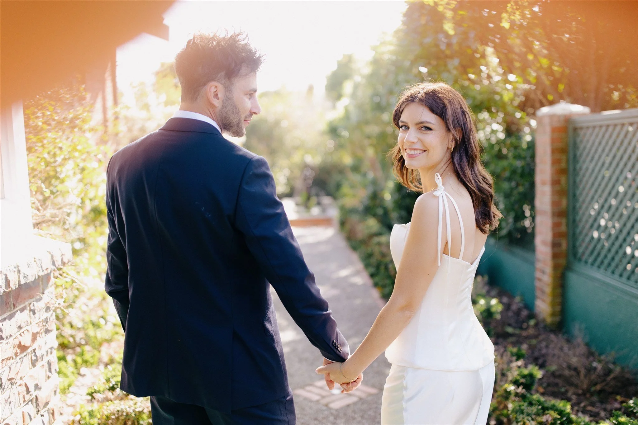 A smiling woman in a white dress holding hands with a man in a dark suit, outside on a sunny day, with greenery and a brick wall in the background.