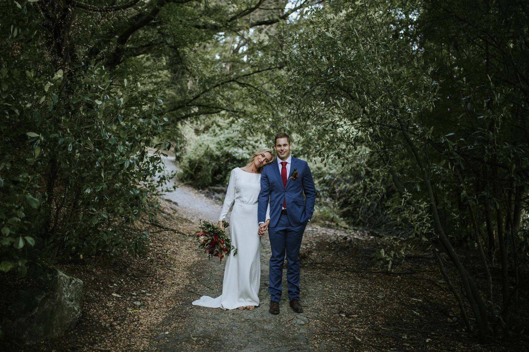 Bride and groom holding hands and smiling on a forested path. The bride wears a white dress and holding a bouquet of red flowers. The groom wears a blue suit with a red tie.