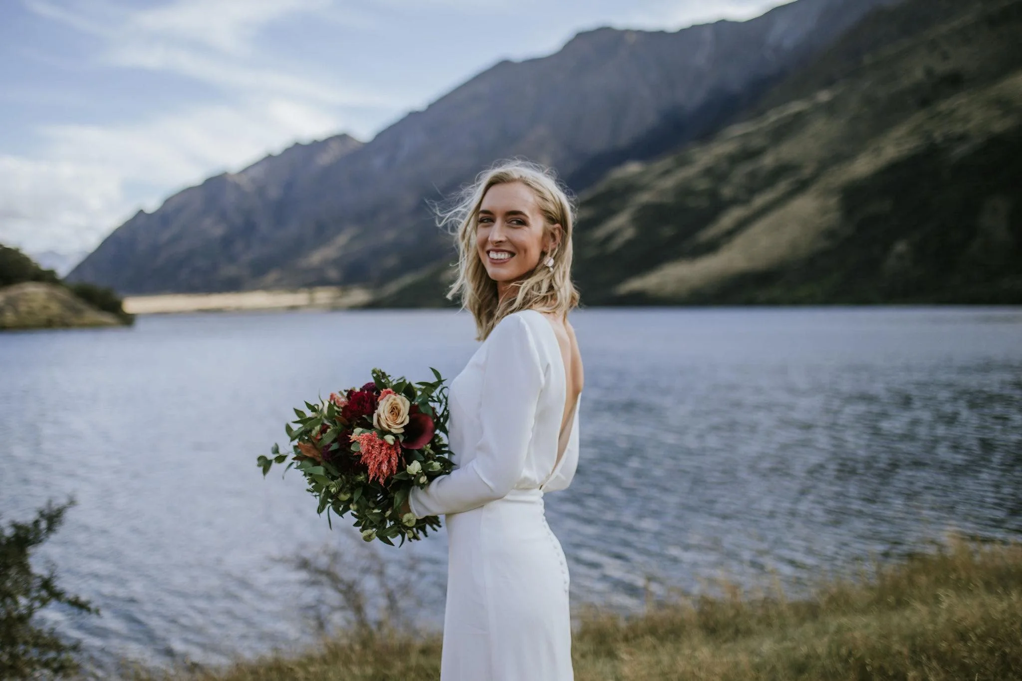 A smiling woman in a white dress holding a bouquet of flowers stands outdoors near a lake with mountains in the background.