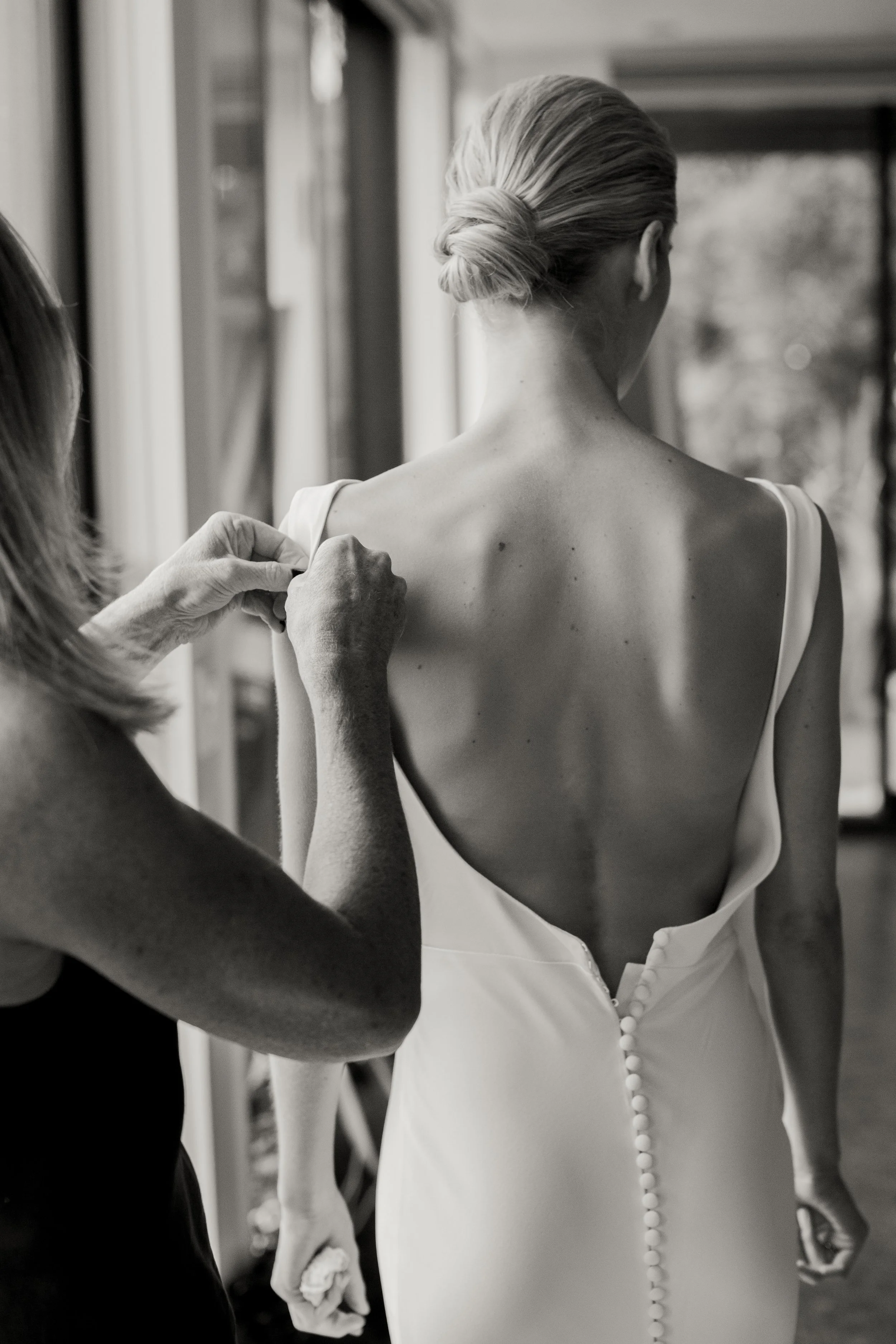 A woman with her back to the camera wearing a wedding dress with buttons down the back, assisted by another woman.
