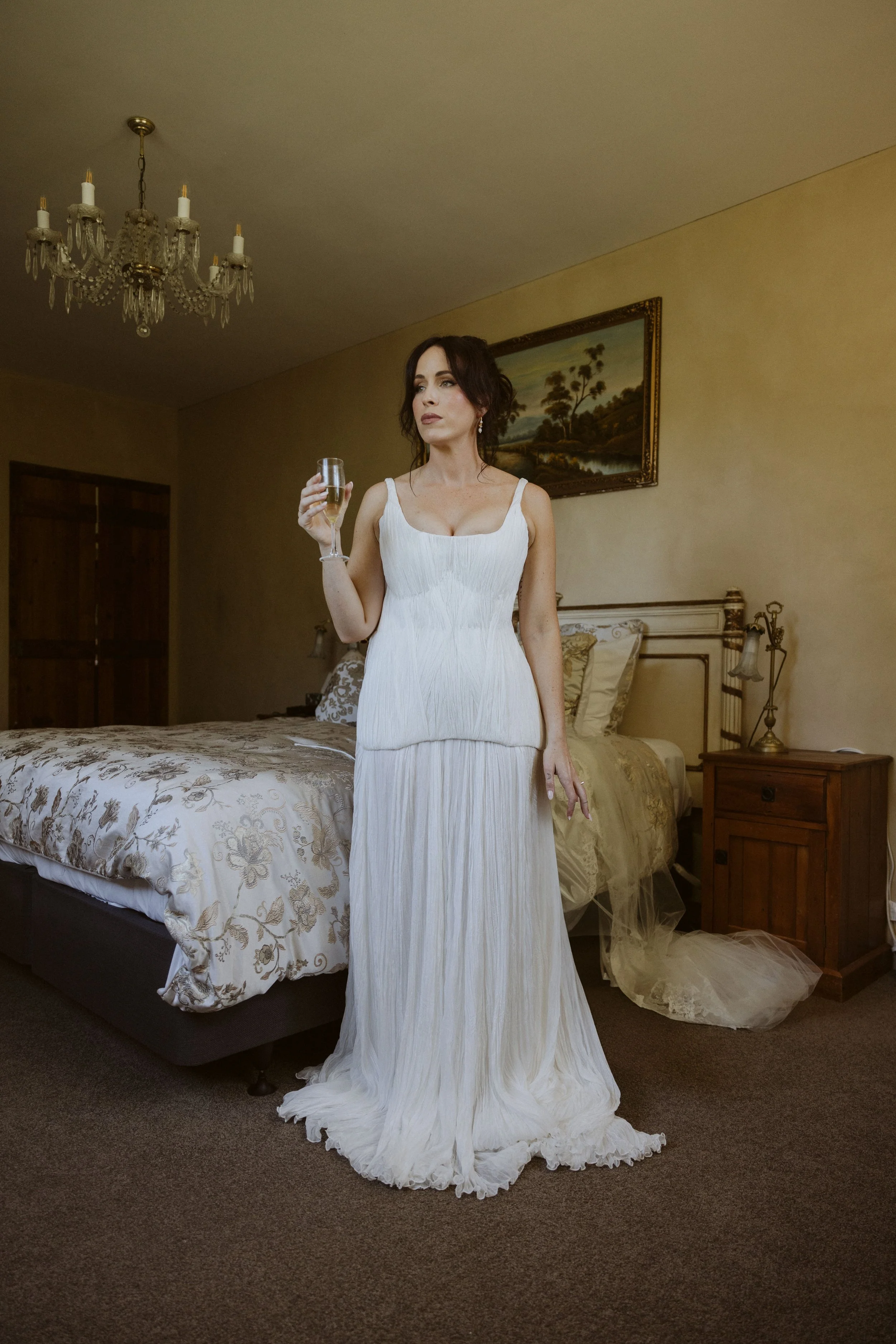 A woman in a white dress standing in a bedroom, holding a glass of wine, with a bed, a painting, and a chandelier in the background.