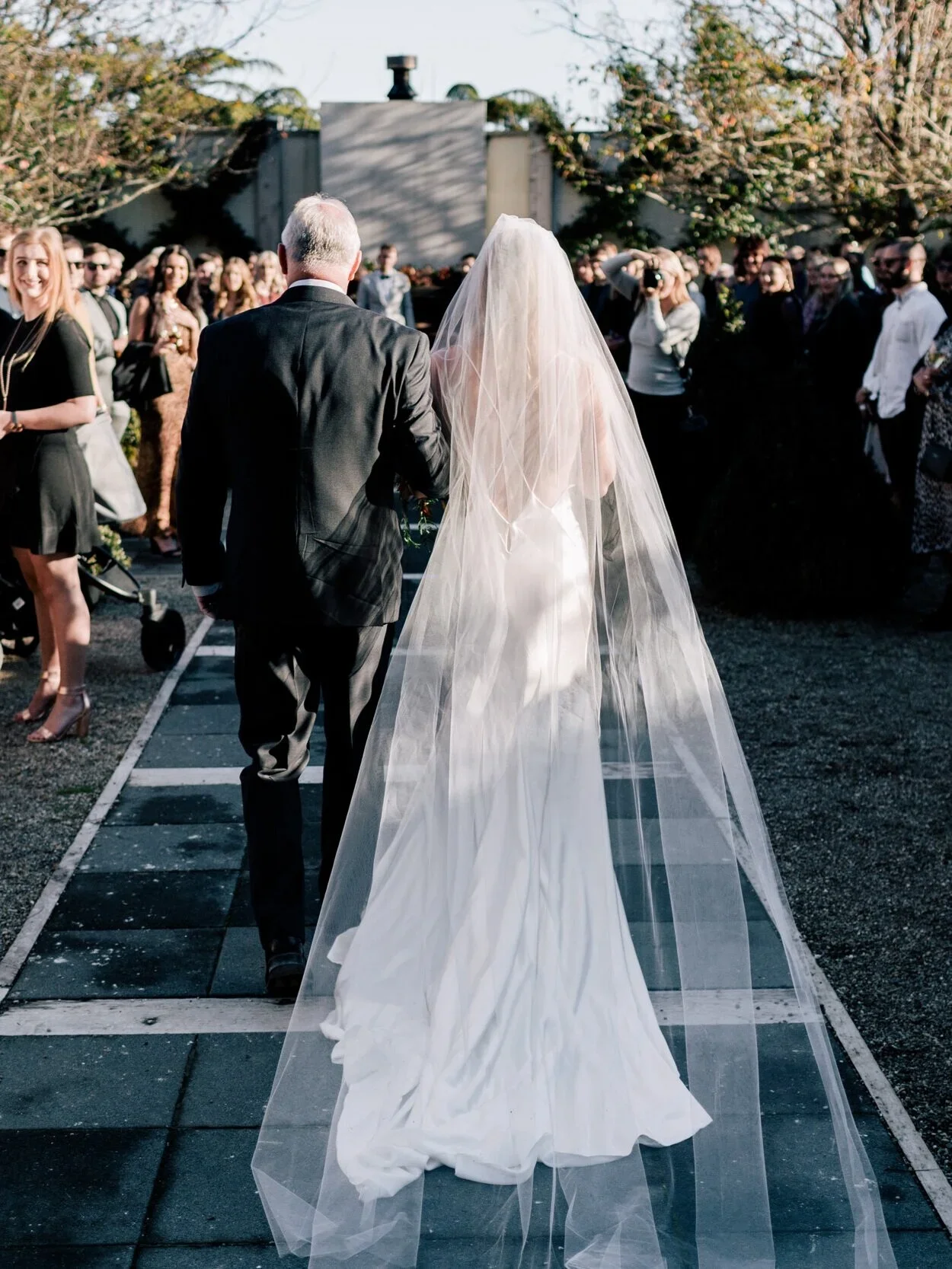 Bride in a long white wedding gown and veil walking down the aisle with her father, surrounded by wedding guests outdoors.