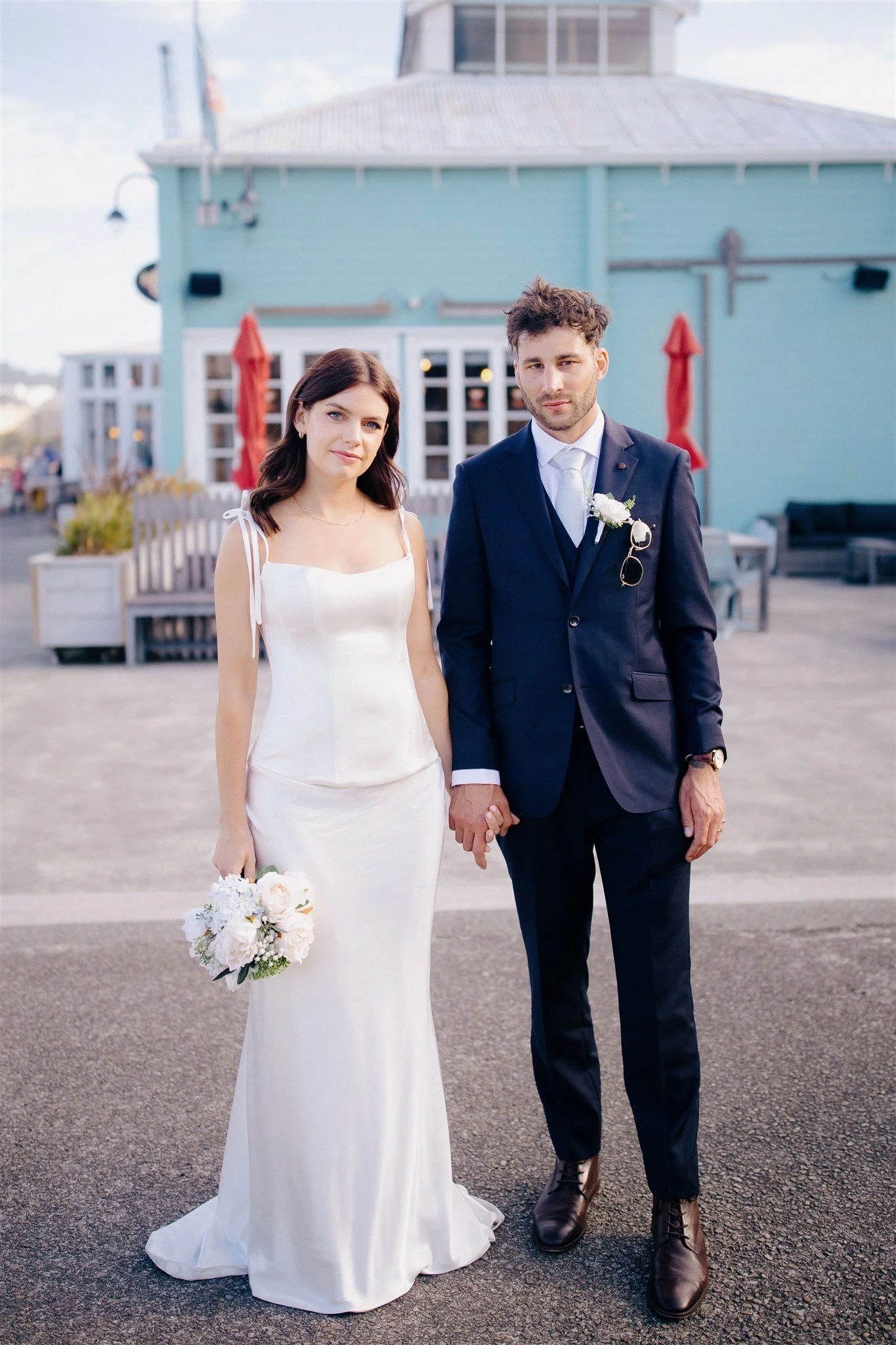 A bride and groom holding hands outdoors, standing on a paved surface in front of a blue building with red umbrellas, during daytime.