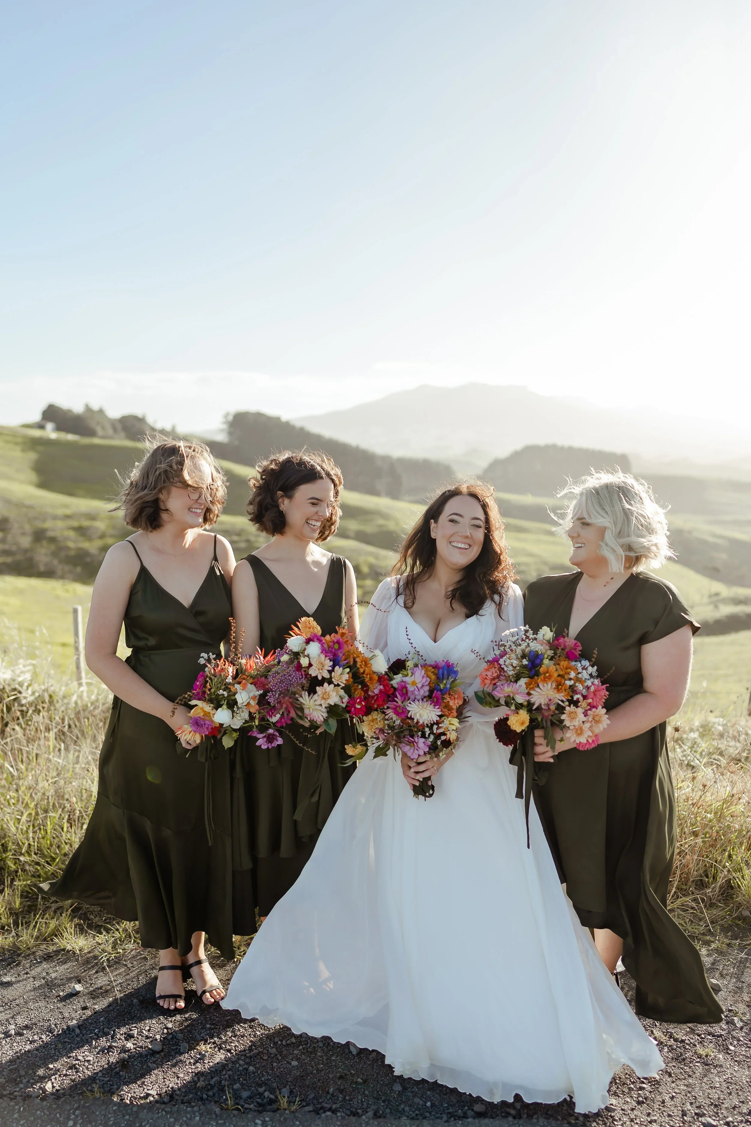 A bride and her three bridesmaids posing outdoors with rolling green hills and mountains in the background, all smiling and holding colorful bouquets.