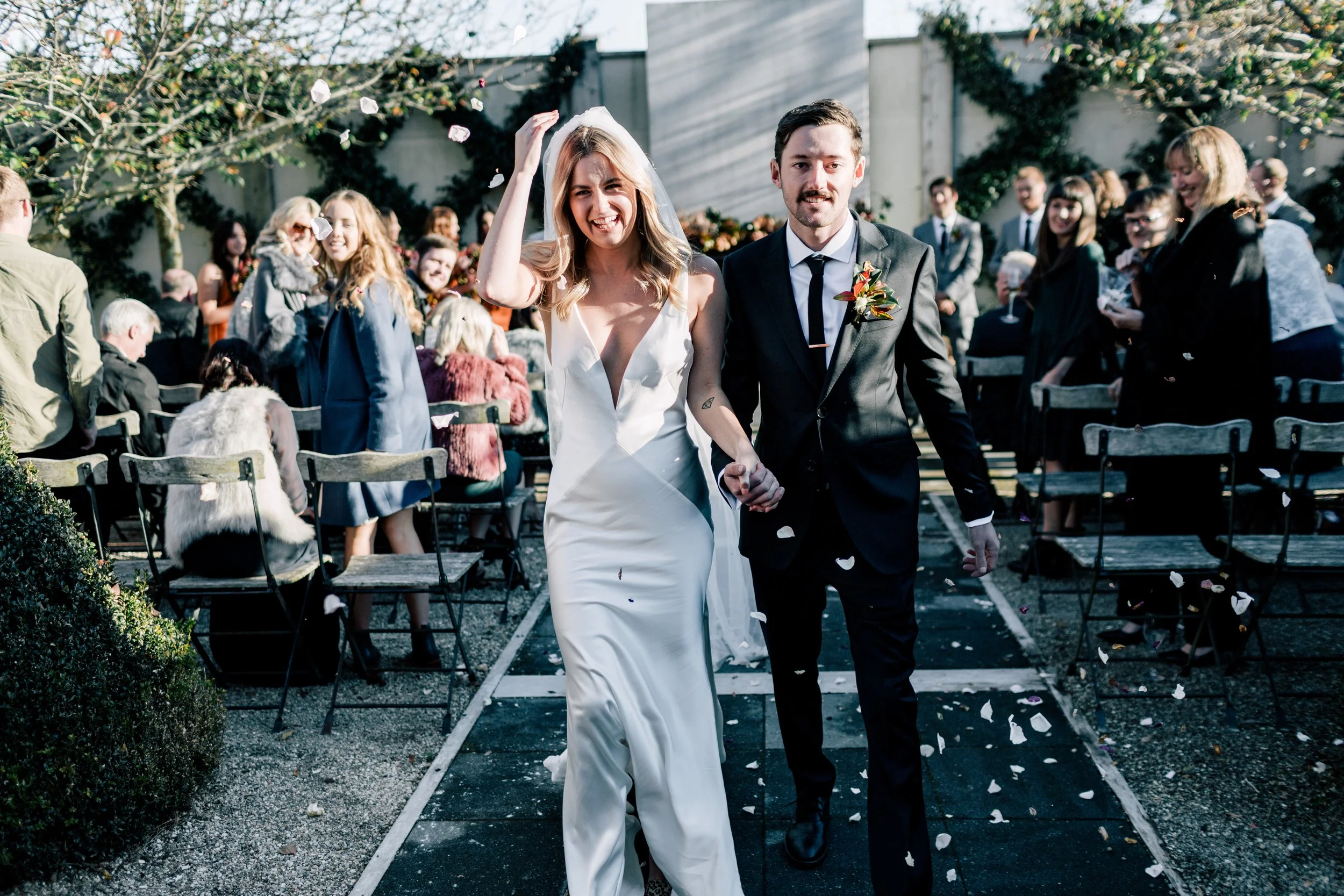 A newlywed couple walking hand in hand down the aisle after their wedding ceremony, surrounded by friends and family, with flower petals falling around them.