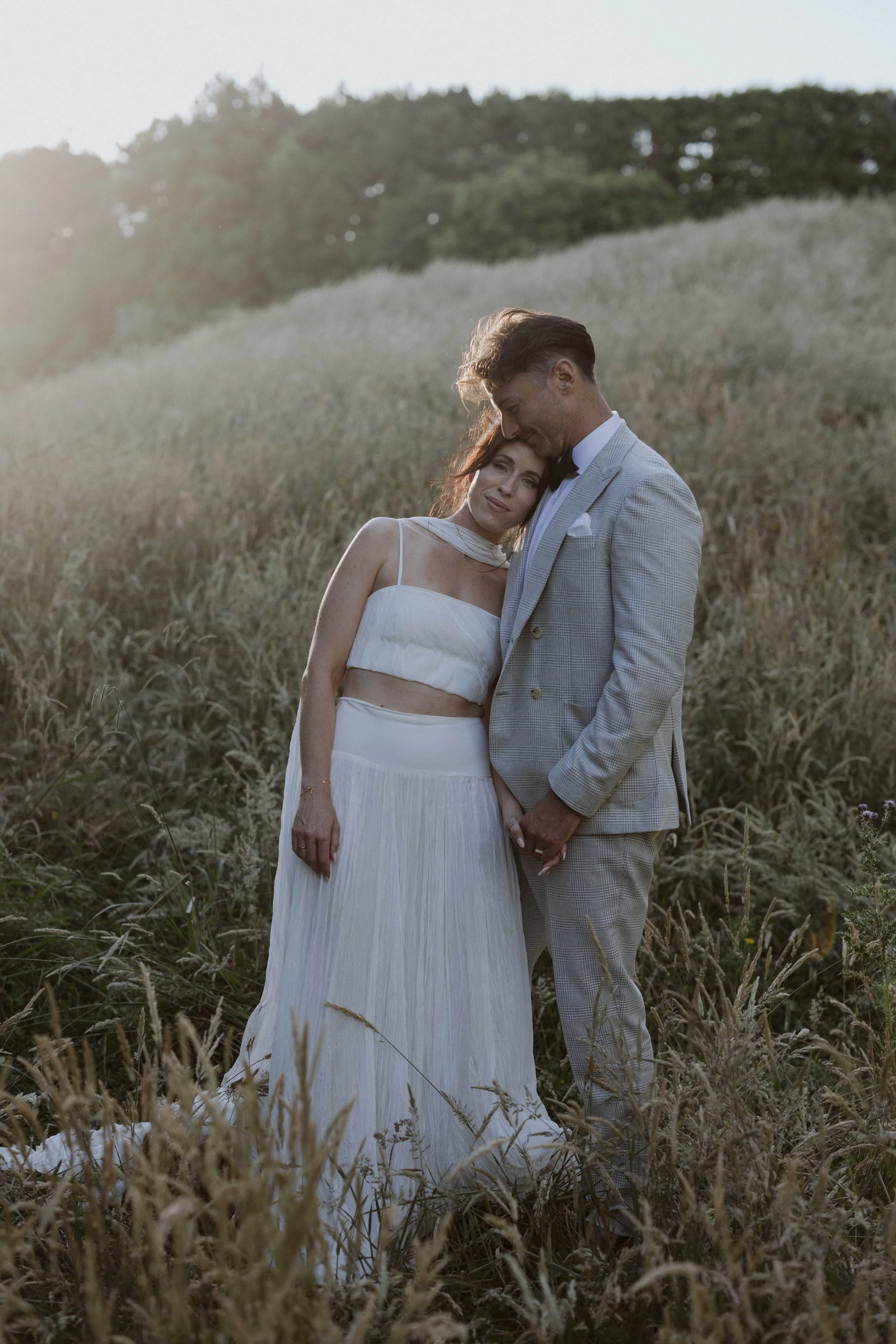 A couple dressed in wedding attire standing together in a field of tall grass, with the woman resting her head on the man's shoulder and holding hands, under soft sunlight.