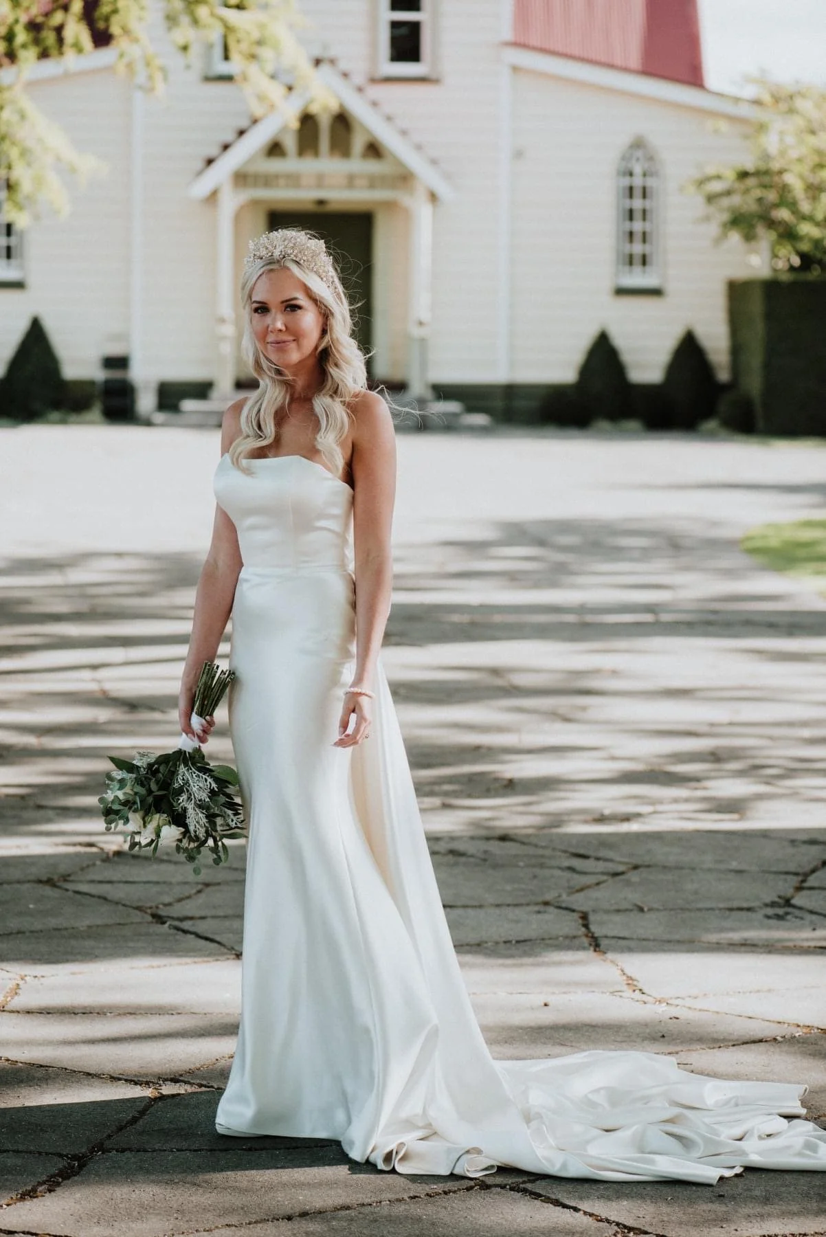 A bride stands outdoors in front of a house, wearing a white wedding gown, holding a bouquet, and wearing a tiara, with trees and shadows in the background.