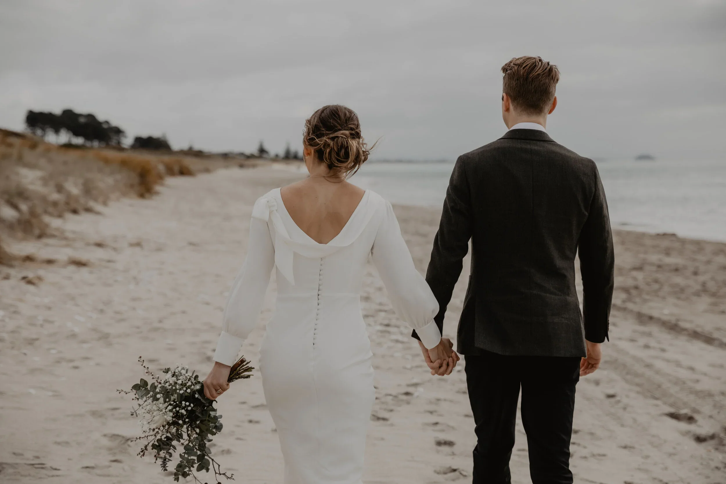 A newlywed couple dressed in wedding attire walking hand in hand along a beach on a cloudy day.