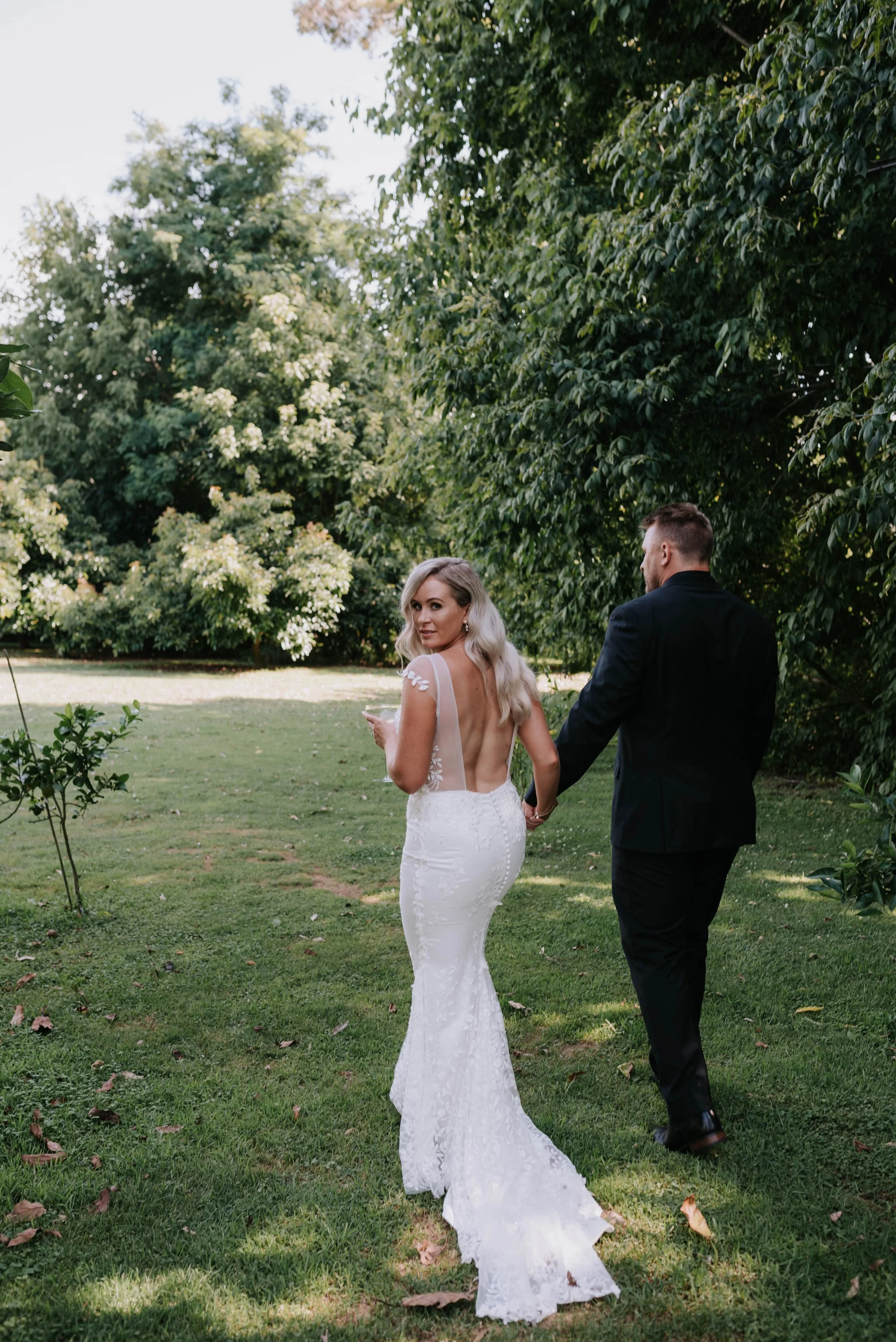 A bride and groom walking hand in hand outdoors on a wedding day in a lush green garden, with the bride glancing back over her shoulder.