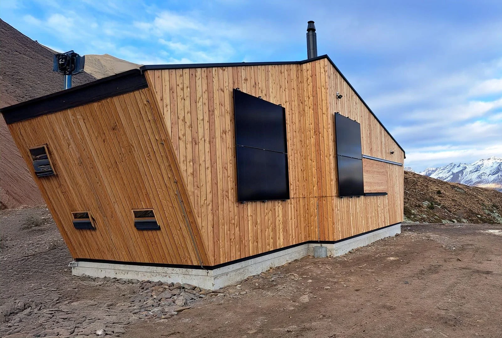 Edificio moderno in legno con grandi pannelli neri, situato in un paesaggio montano con neve sullo sfondo.