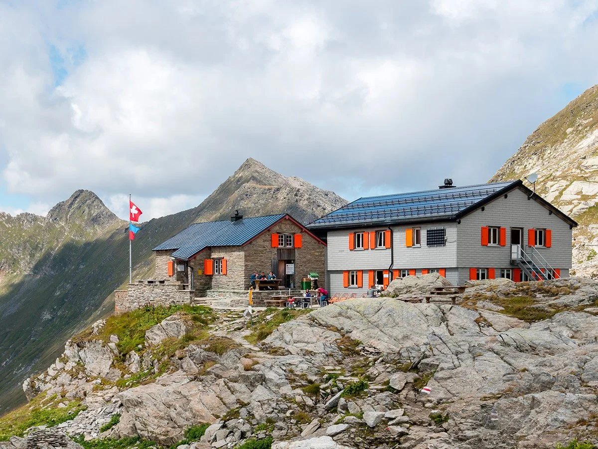 Due rifugi di montagna in un paesaggio alpino con rocce, montagne e nuvole, uno con bandiera svizzera.