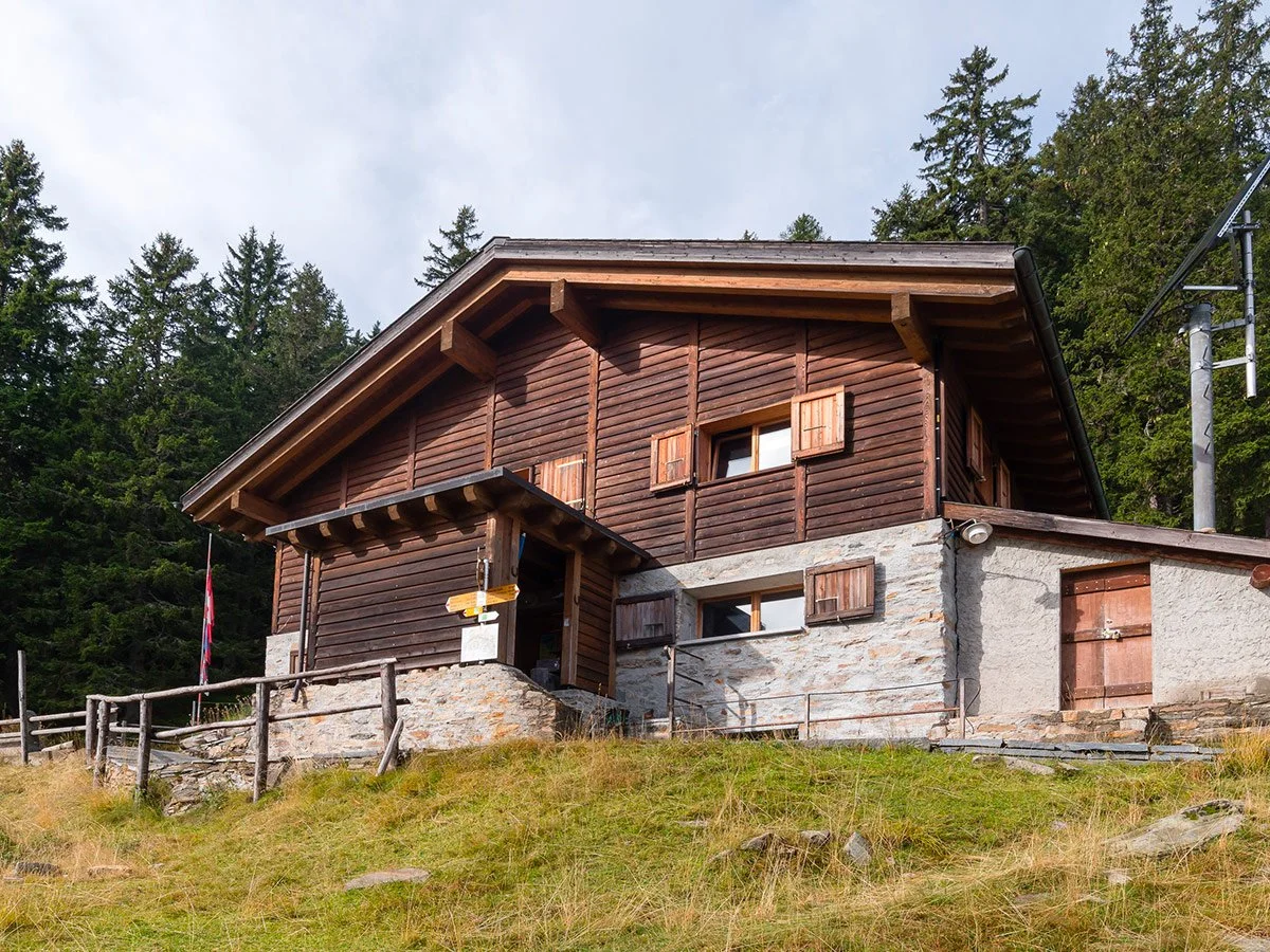 Casa di montagna in stile rustico con muro in pietra e legno, situata su un declivio erboso, con una rete di recinzioni e alberi di conifere sullo sfondo.