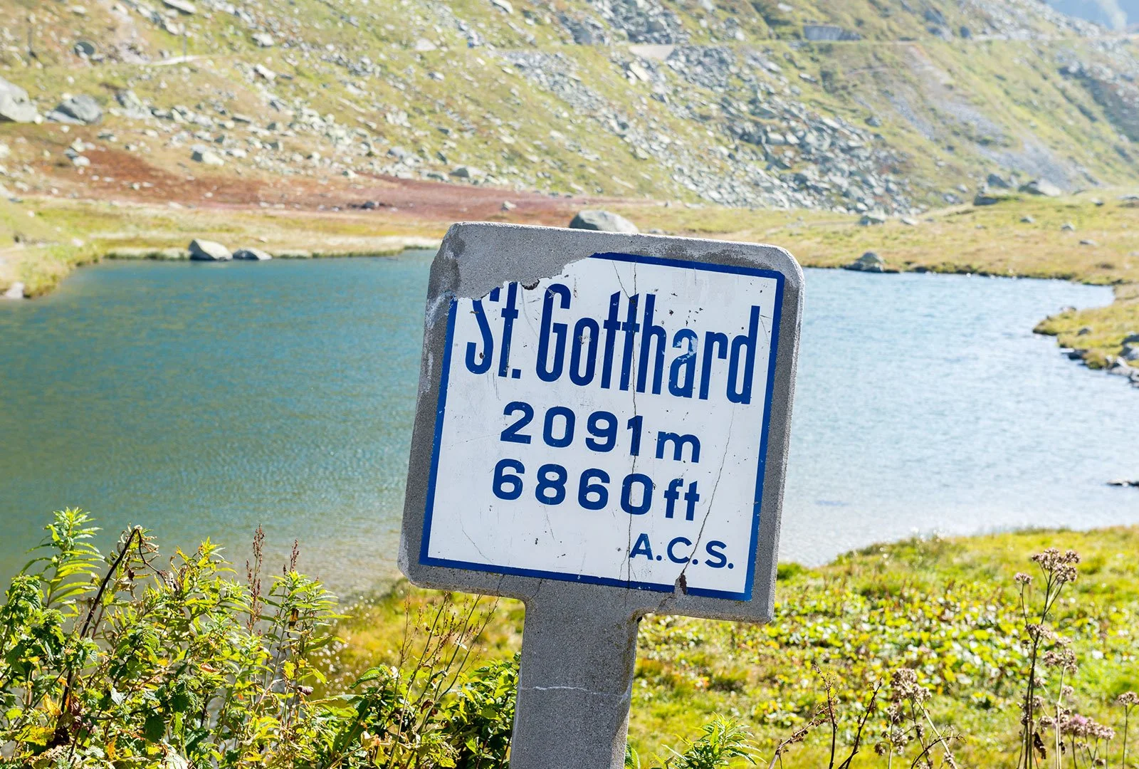 Segnale che indica l'altitudine di St. Gotthard, 2091 metri o 6860 piedi, in un paesaggio montano con un lago e vegetazione di montagna.