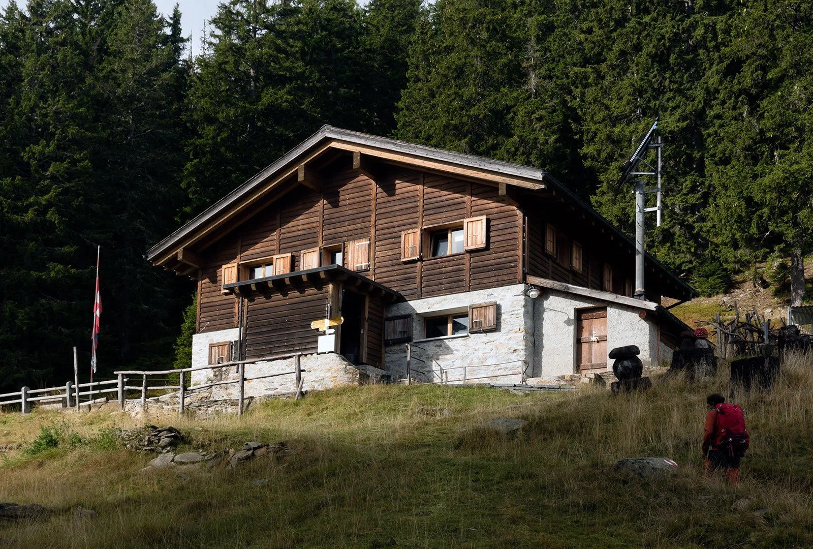 Una casa di montagna in stile tradizionale circondata da alberi e prati, con una persona che cammina sul terreno in primo piano.