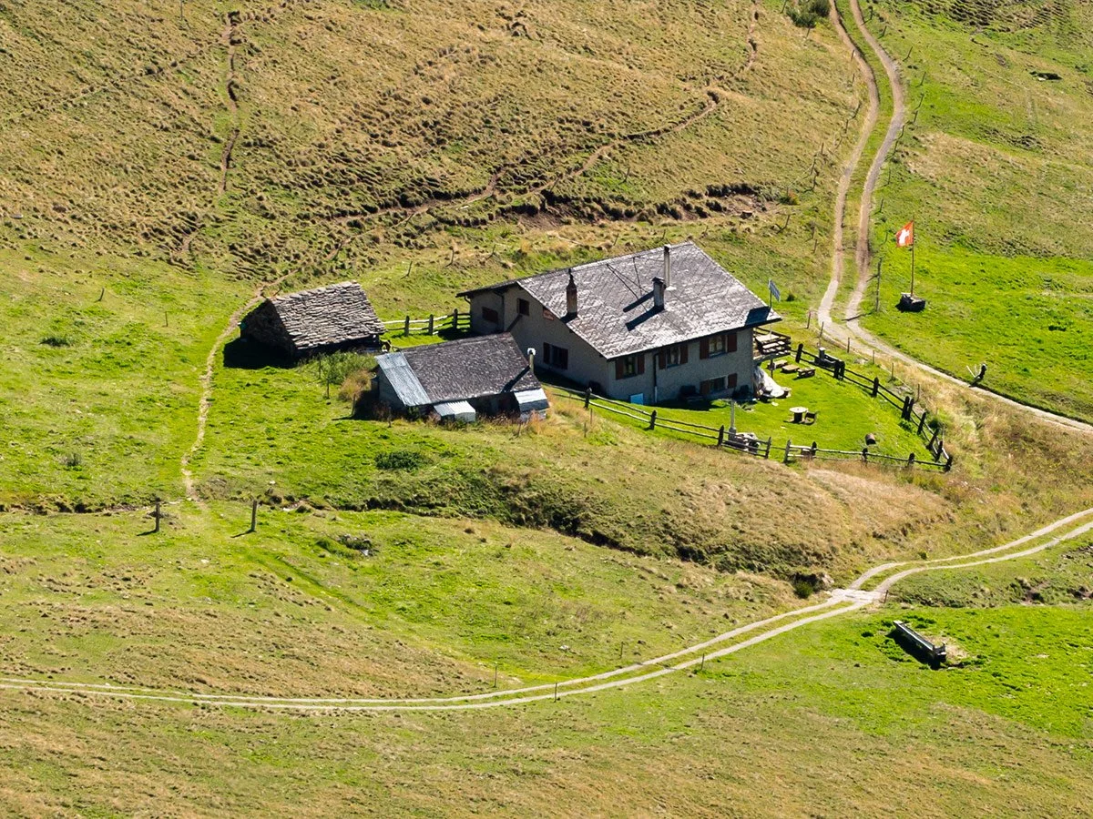 Casa di montagna circondata da campi verdi, con sentieri e un'area recintata con sedie e tavoli, bandiera svizzera nel prato, strada sterrata vicina.