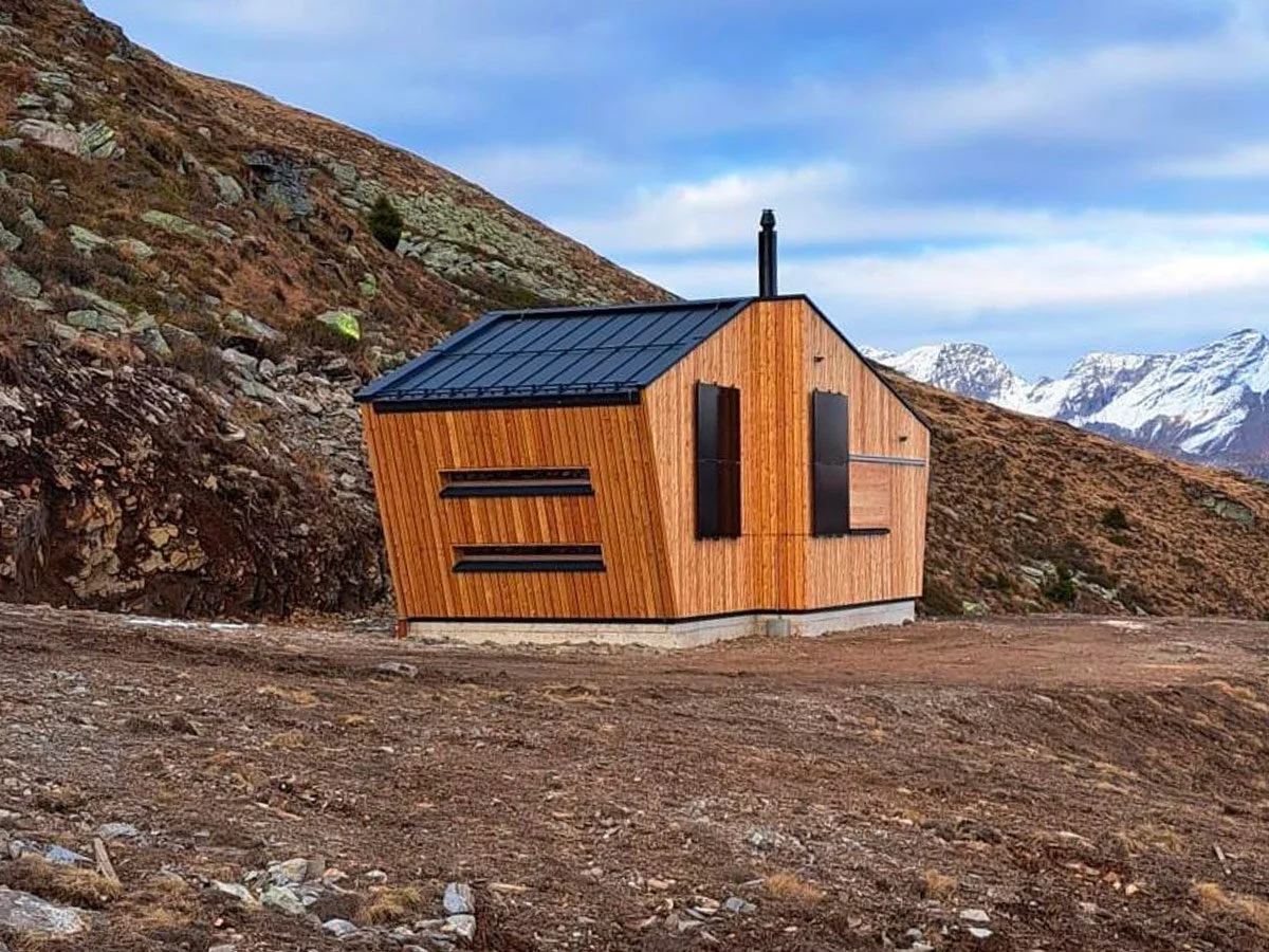 Una casetta moderna in legno su un terreno montano, con montagne innevate sullo sfondo.