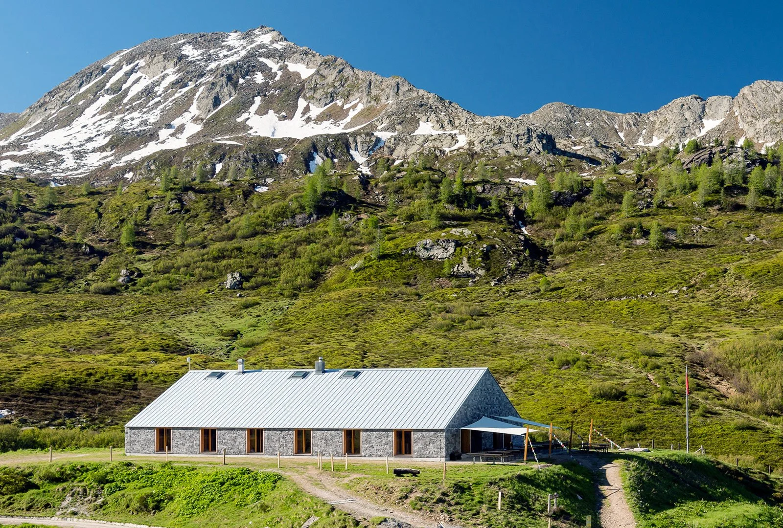 Una casa di pietra con un tetto in metallo, situata in un paesaggio montano verde con neve sulle cime, sotto un cielo blu.