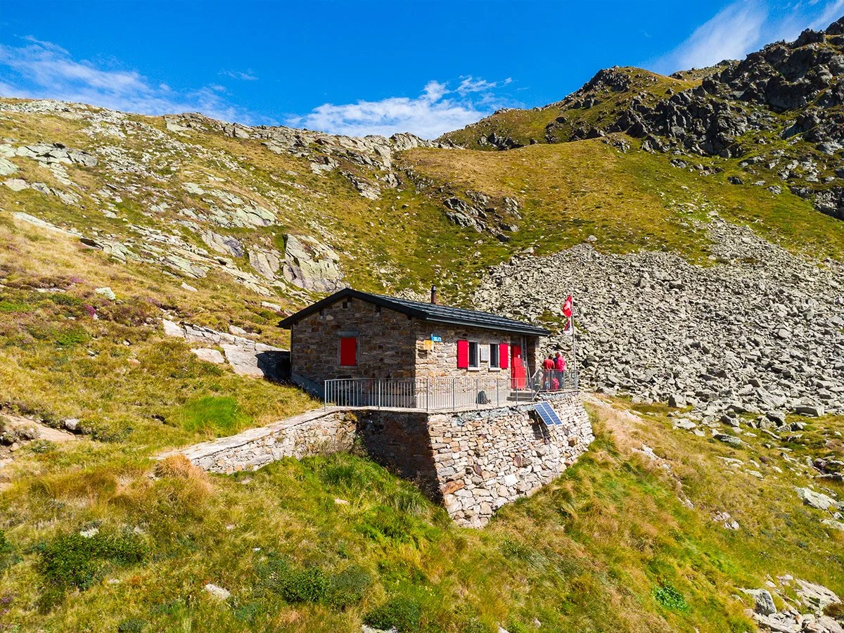 Casa di pietra con tetto di tegole in montagna, bandiera svizzera accanto e pannello solare in giardino, circondata da erba e rocce, sotto cielo blu con alcune nuvole.