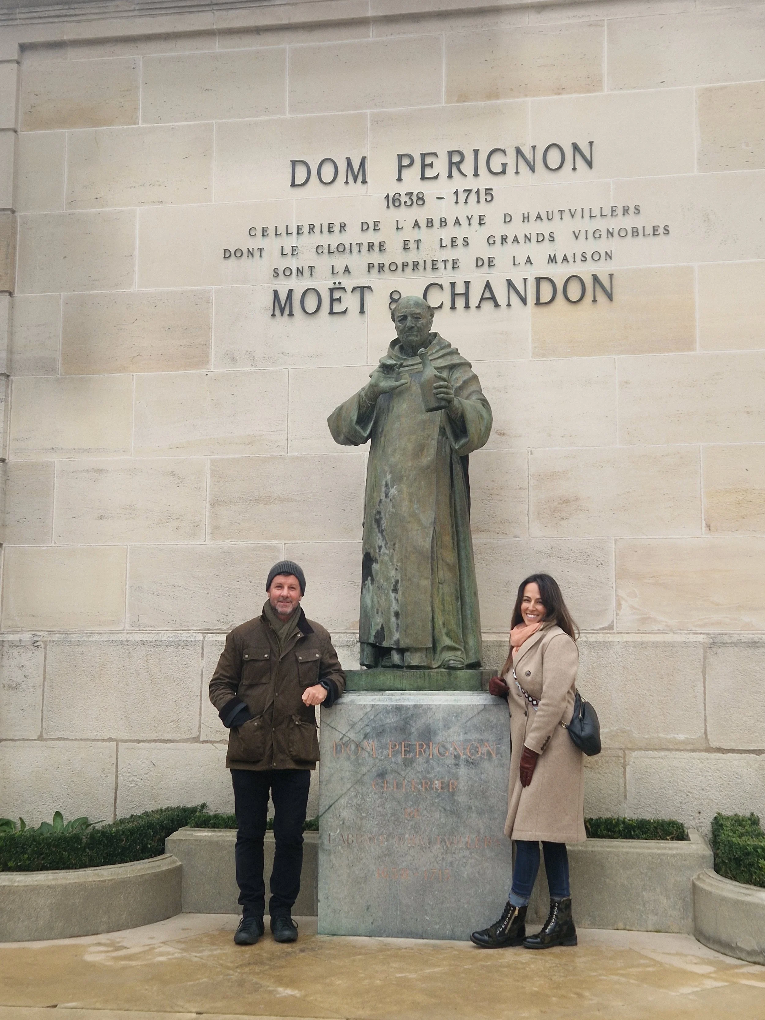 A man and woman standing next to a large statue of Dom Perignon in front of a wall with French text, both smiling and dressed warmly.