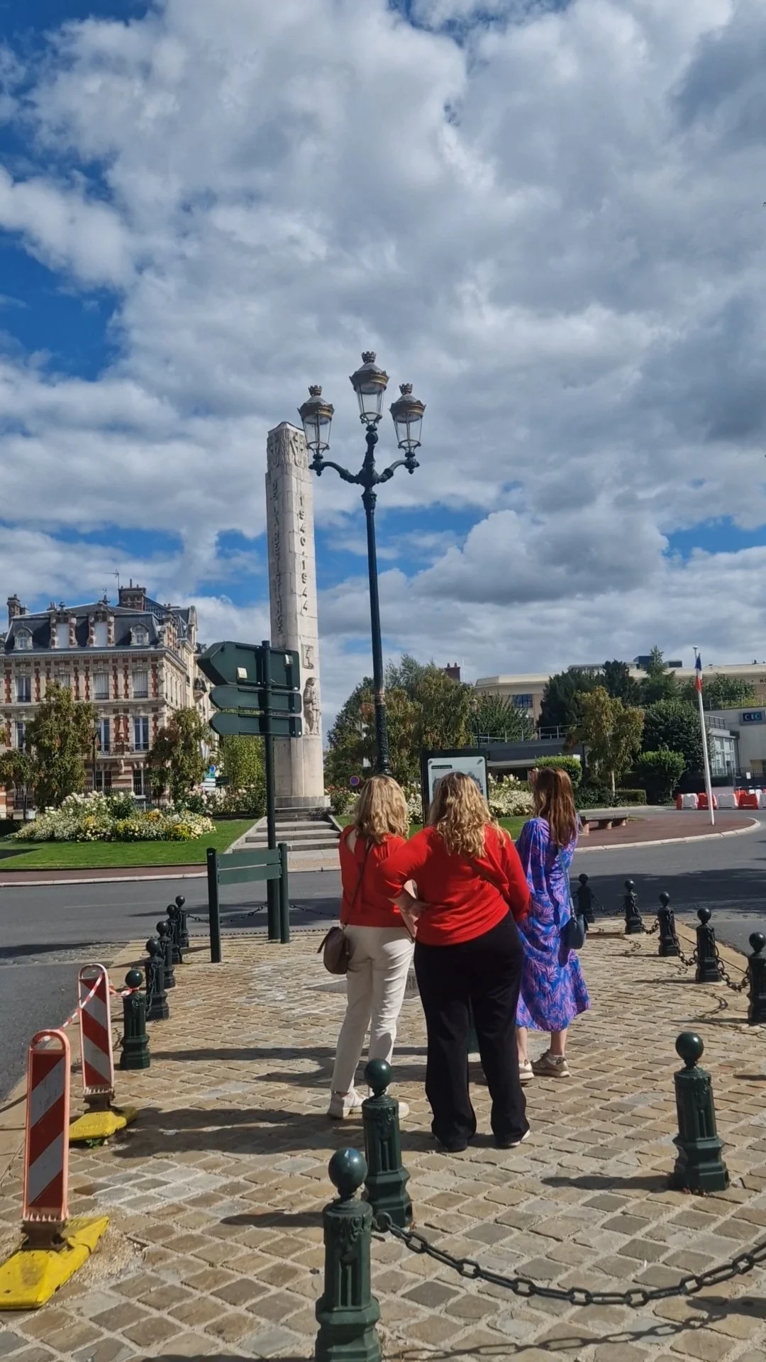 Three women standing on a cobblestone plaza, tourists in a city with trees, building, a monument with an inscription, a black lamp post, and a French flag in the background.