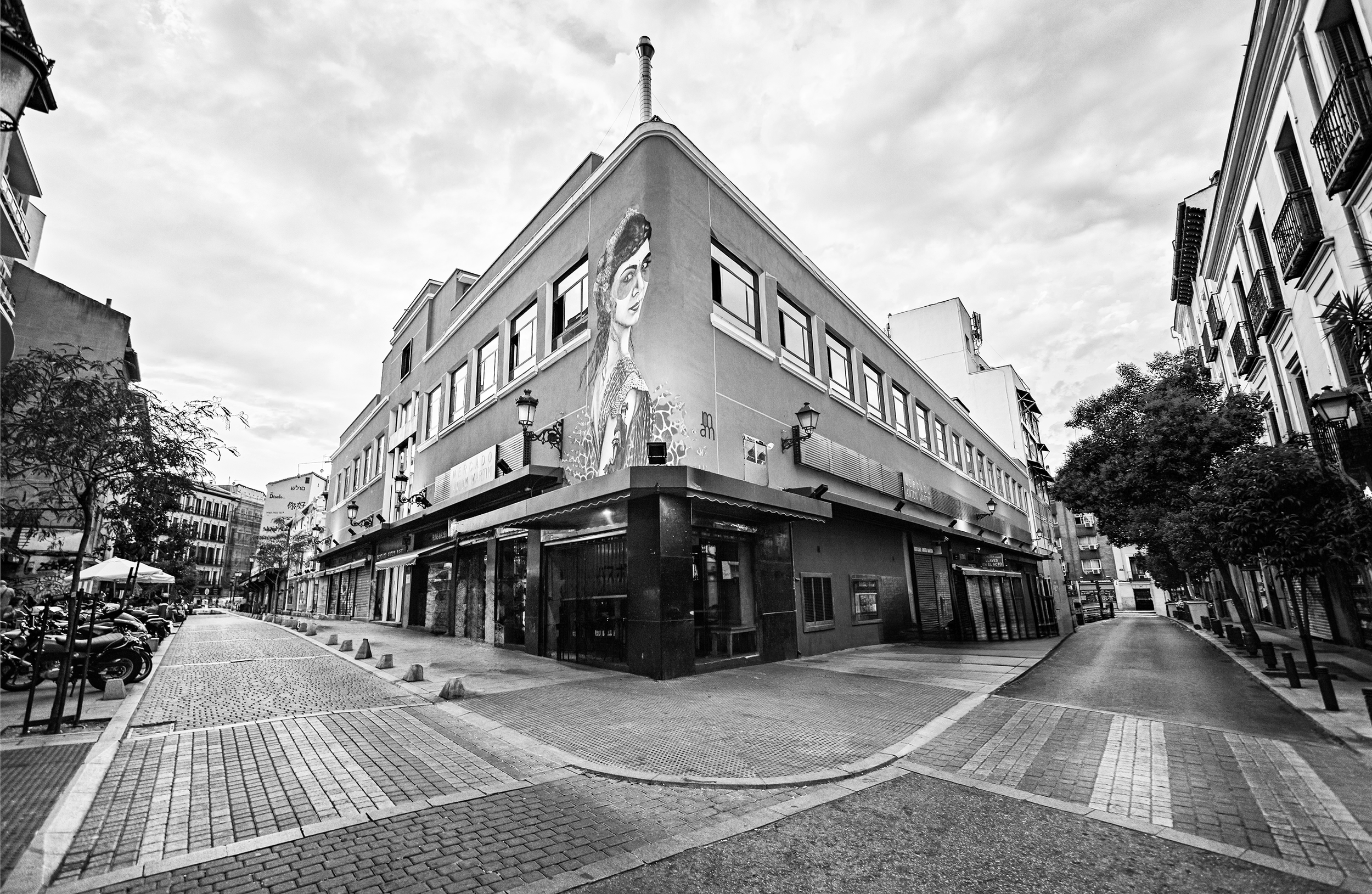 Vista de una calle en una ciudad con edificios, en la esquina de un edificio hay un mural de una mujer con gafas y peinado recogido. La calle es peatonal con algunos automóviles y motocicletas estacionados. La imagen es en blanco y negro.