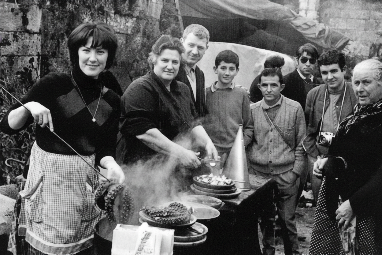 Grupo de personas alrededor de una mesa con comida en un entorno exterior, en una fotografía en blanco y negro.