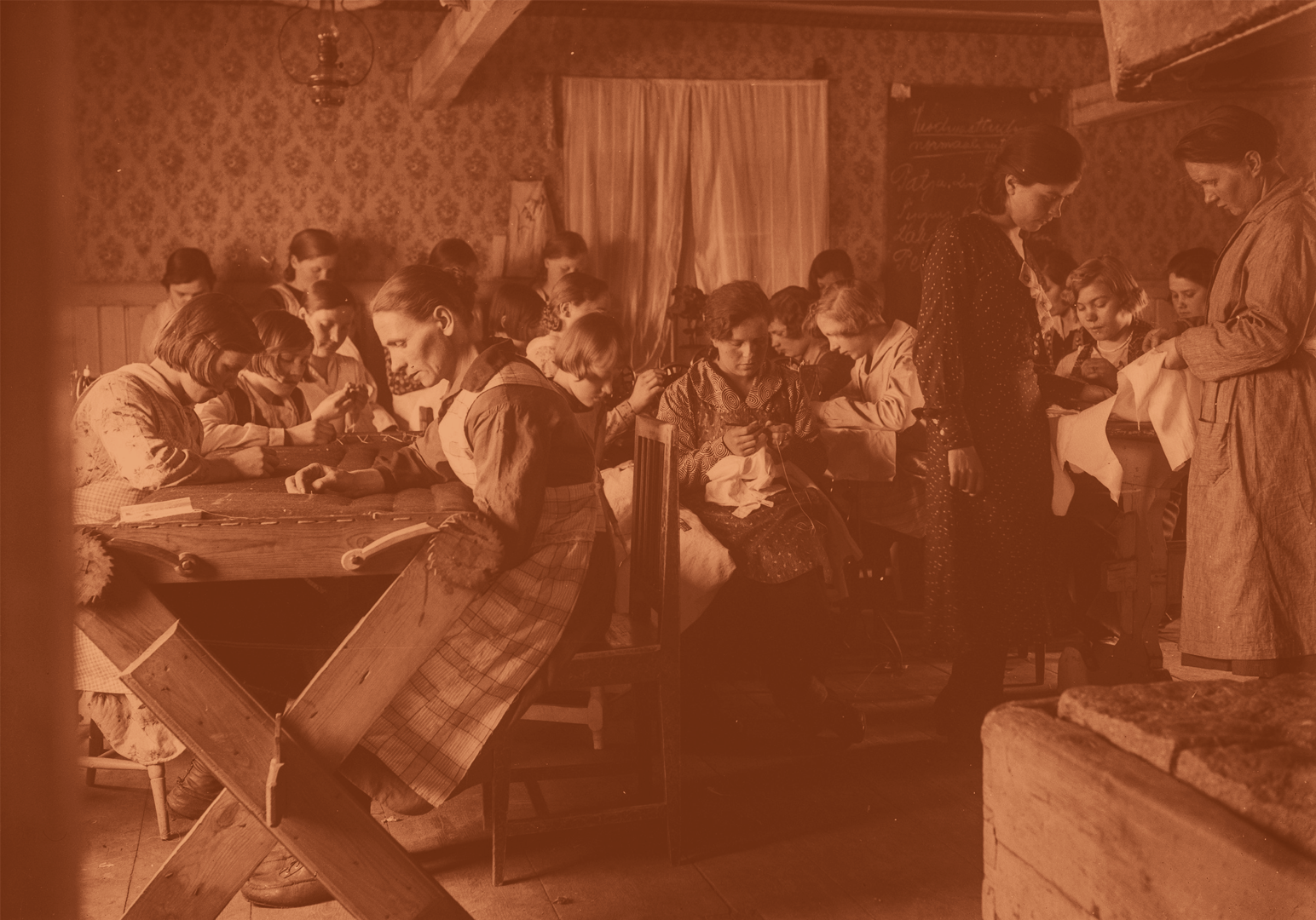 Vintage black-and-white photo of women and children sitting at tables engaged in needlework inside a cozy room with floral wallpaper and curtains.