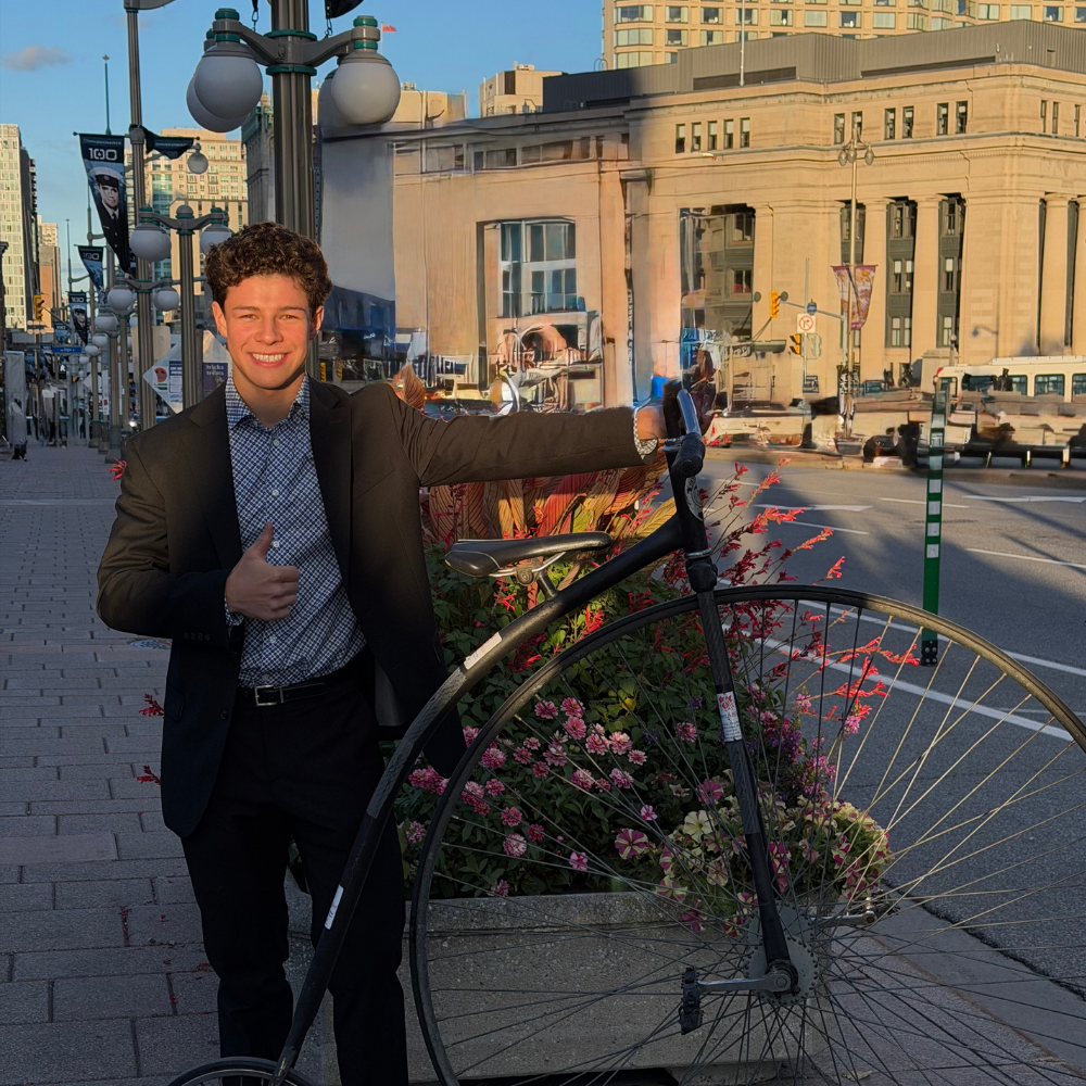 A young man in a black suit smiling and giving a thumbs-up with one hand, standing on a city sidewalk next to a vintage penny-farthing bicycle and a flower planter. The cityscape with buildings and cars is visible in the background.