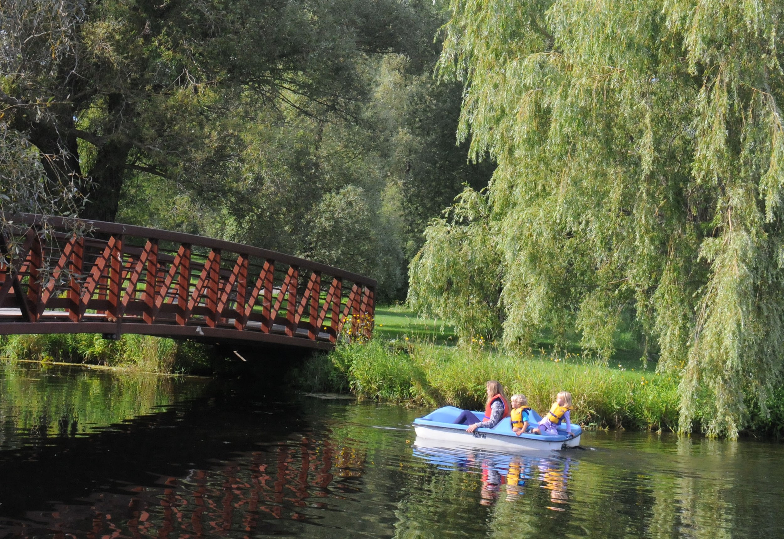 A woman and two children in a blue paddle boat on a calm river, surrounded by lush green trees and a small wooden bridge over the water.