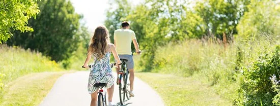 A girl and a boy riding bicycles on a sunny countryside trail surrounded by green trees and grass.