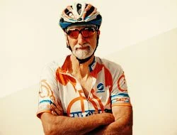 Older man wearing sunglasses, a cycling helmet, and a colorful cycling jersey with short sleeves, standing against a plain background.