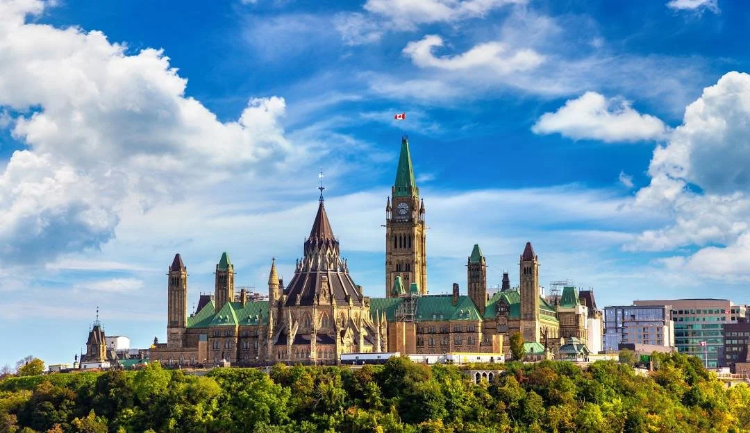 Ottawa Parliament Hill with the Peace Tower, central building, and surrounding structures under a partly cloudy sky, with green trees in the foreground.