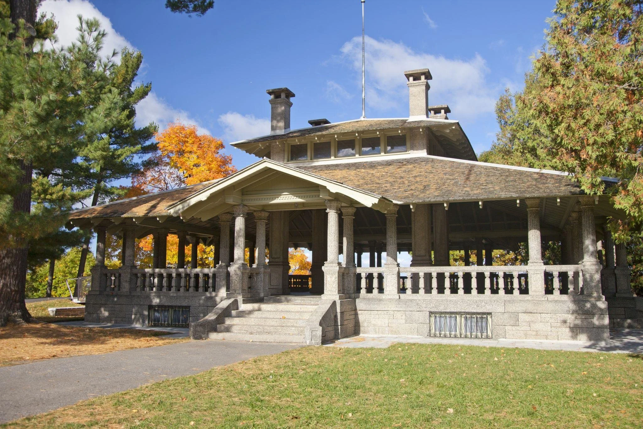 A stone pavilion with a wrap-around porch and multiple stone columns, set in a park with autumn foliage and a clear blue sky.
