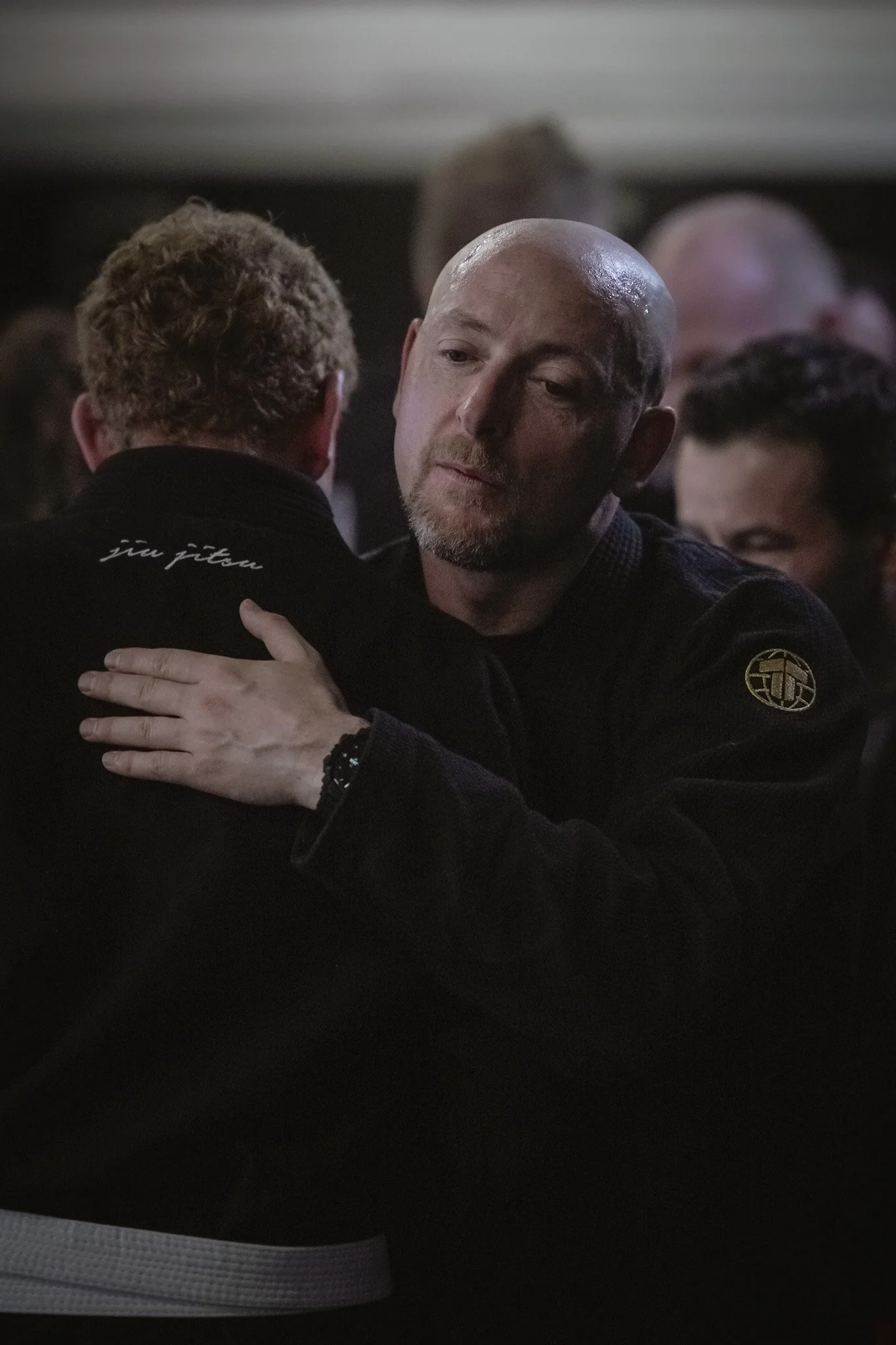 Two men hugging each other at a martial arts event, with the man facing the camera showing a shaved head and beard.