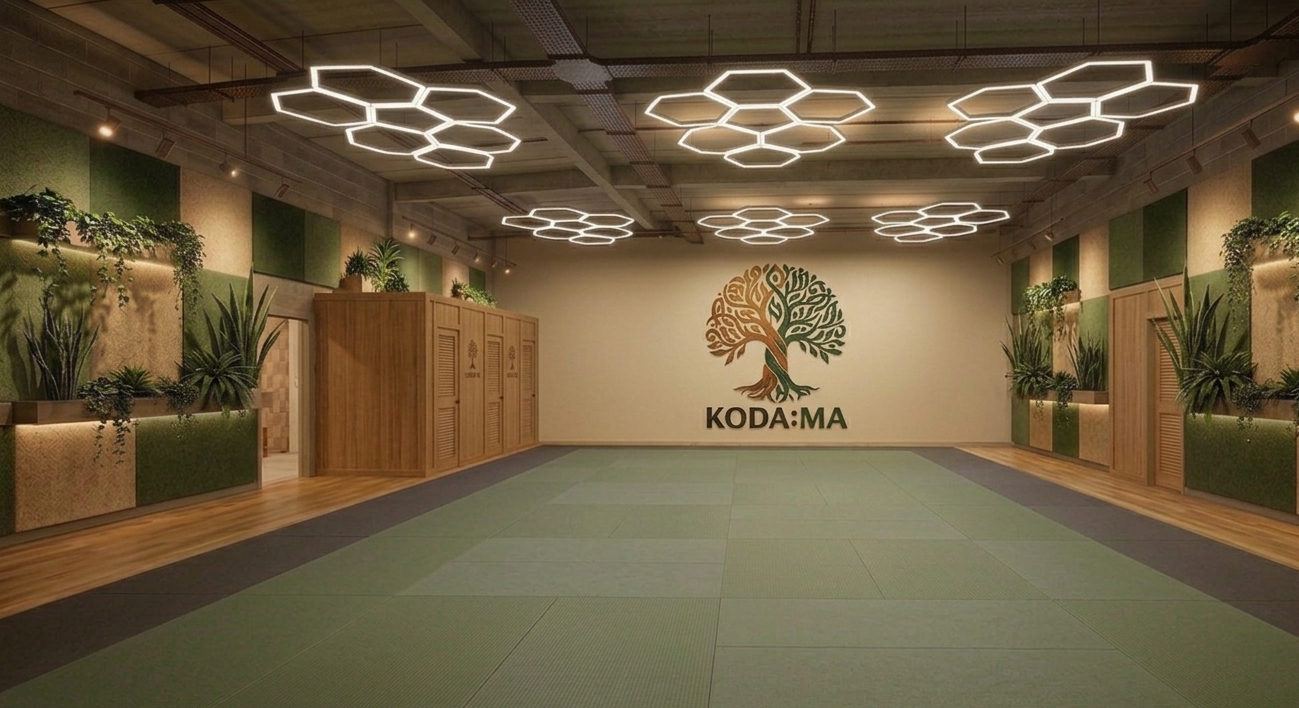 A spacious yoga or martial arts studio with a green mat floor, decorative plants along the walls, wooden storage lockers, and a large wall decal of a tree with the words "KODA:MA" underneath. Modern hexagon-shaped ceiling lights illuminate the space.