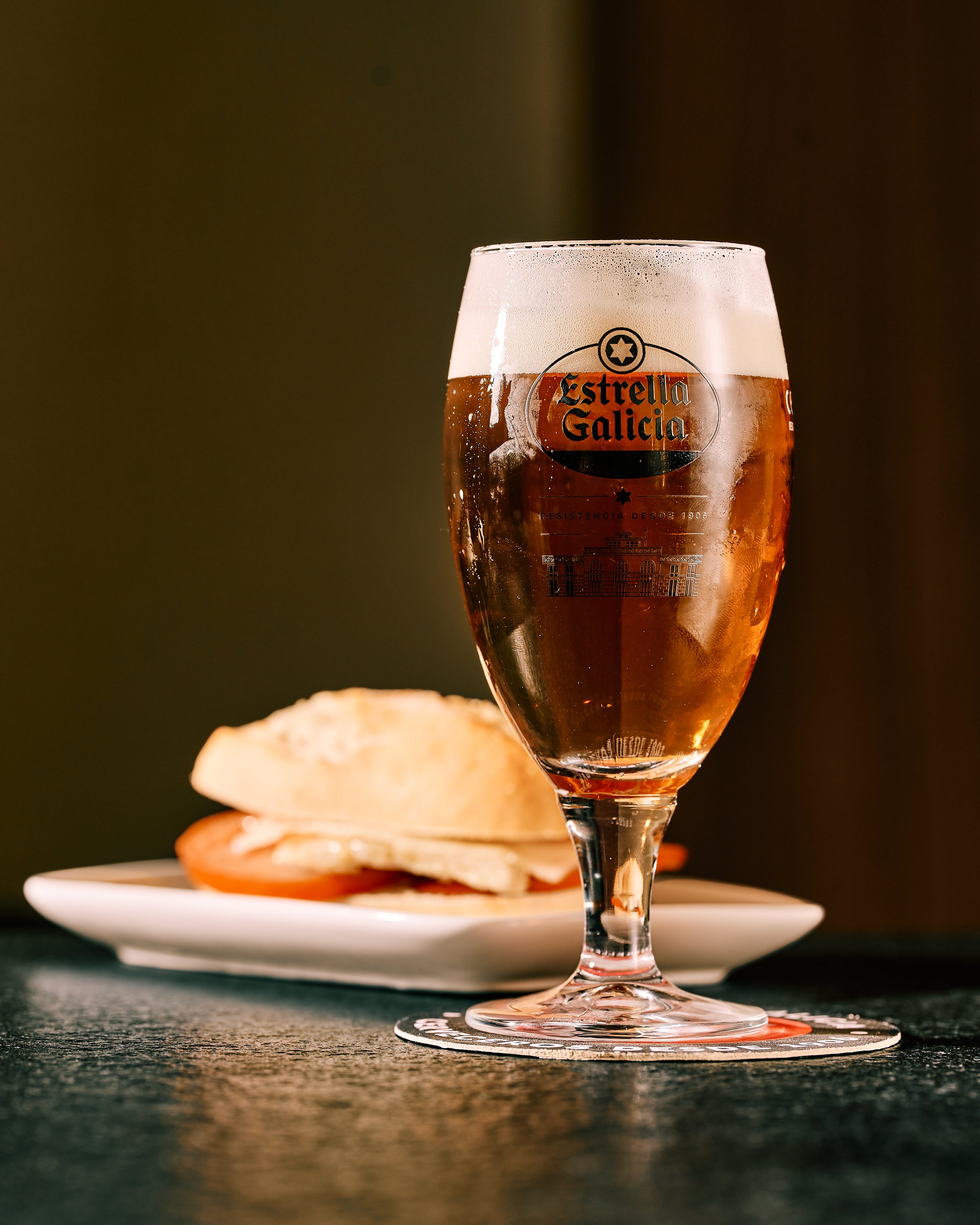 Vaso de cerveza Estrella Galicia con espuma en la parte superior, al fondo un plato con un piripi; pan, tomate, lomo, queso y bacon sobre una mesa oscura.