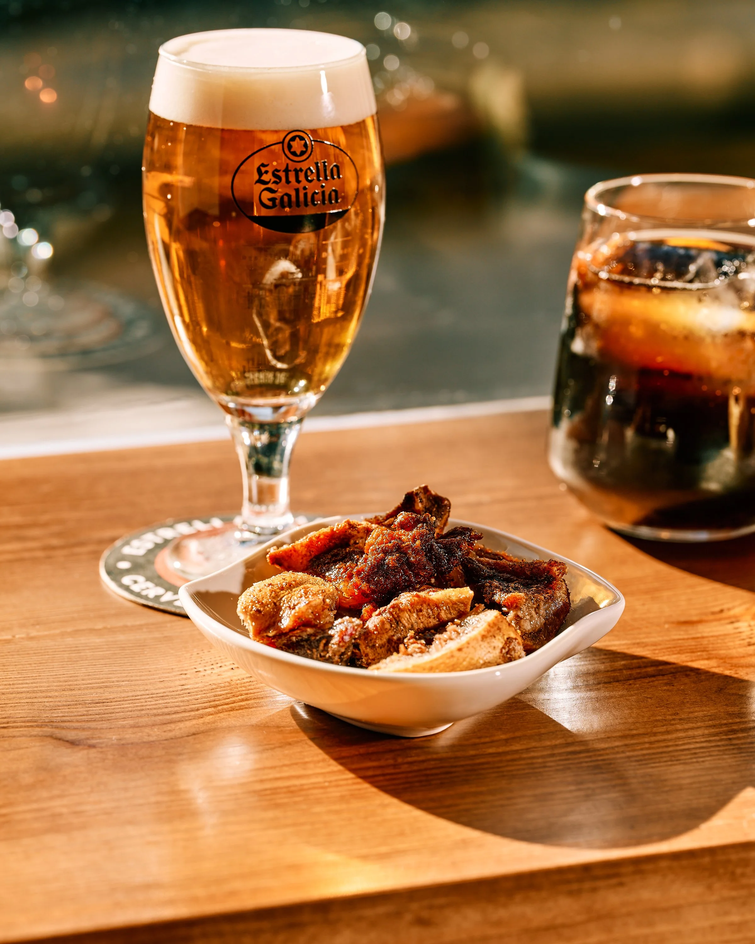 Vaso de cerveza Estrella de Galicia y un vaso de vermú con hielo en una mesa de madera, con un pintxo de jeta