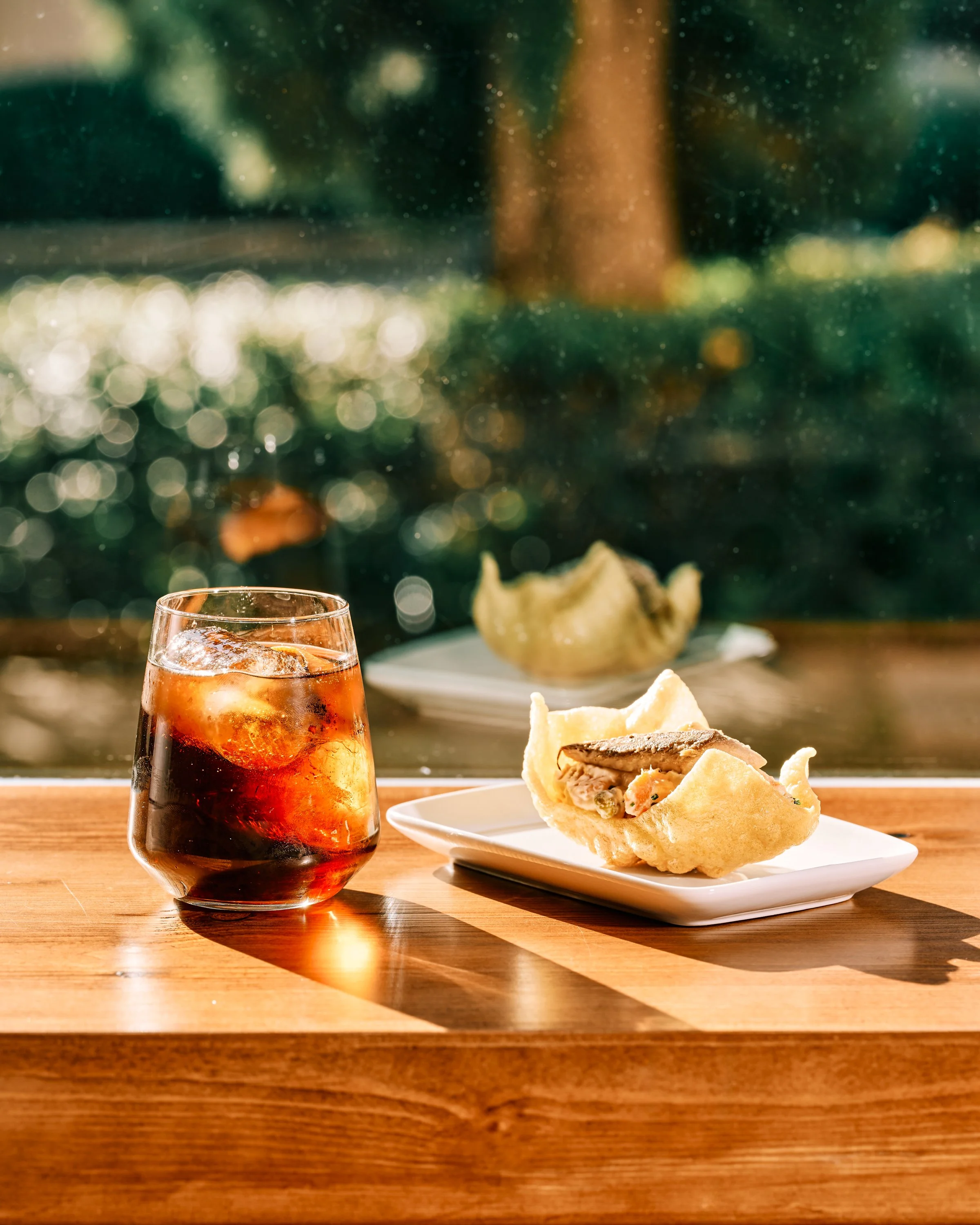Vaso de vidrio con vermú y hielo junto a un plato que tiene un pintxo de paloma en una mesa de madera, con fondo de ventana y naturaleza.