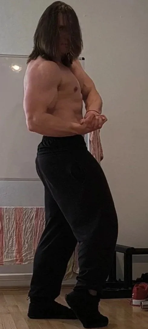 Ein muskulöser Mann mit langen Haaren, der in einem Zimmer neben einem Spiegel steht und seine Armmuskeln zeigt.