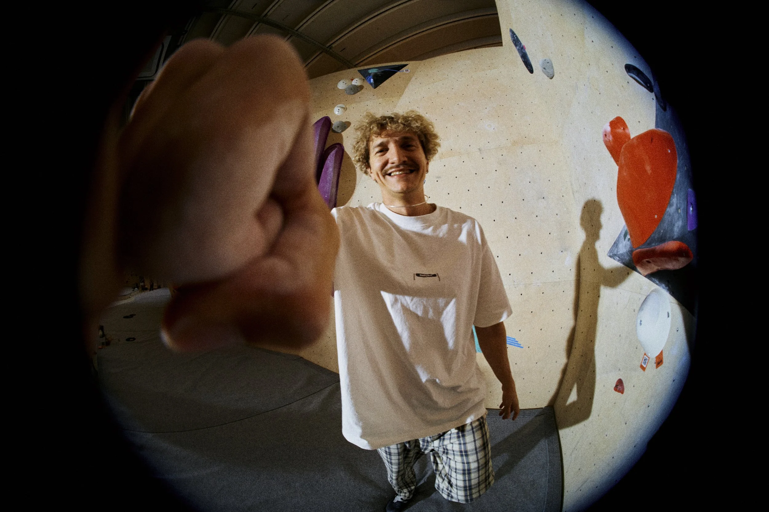 Young man smiling and reaching out, seen through a fisheye lens in an indoor rock climbing gym with climbing holds and a beige climbing wall.