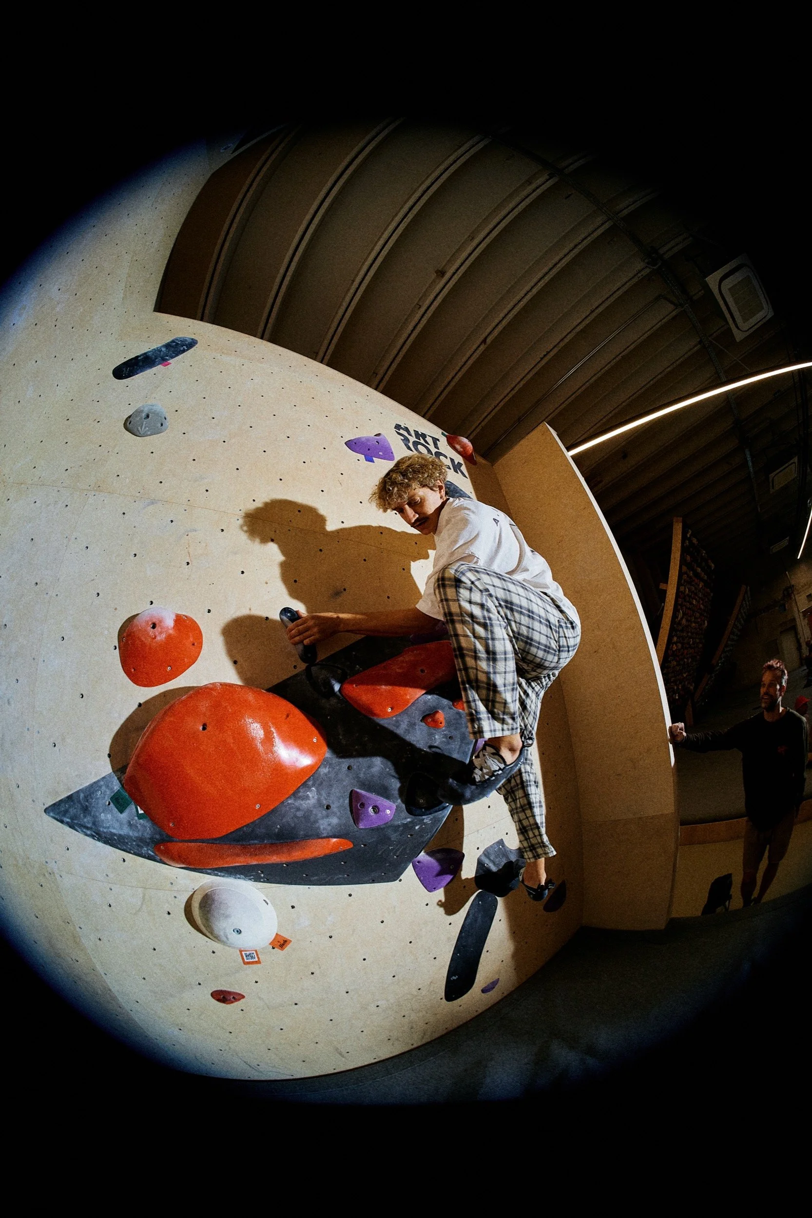 Junge beim Bouldern an einer Kletterwand, trägt karierte Hosen und einen weißen T-Shirt, im Hintergrund eine Person mit dunklem Hemd, umgeben von Klettergriffen und -zubehör in einer Kletterhalle.