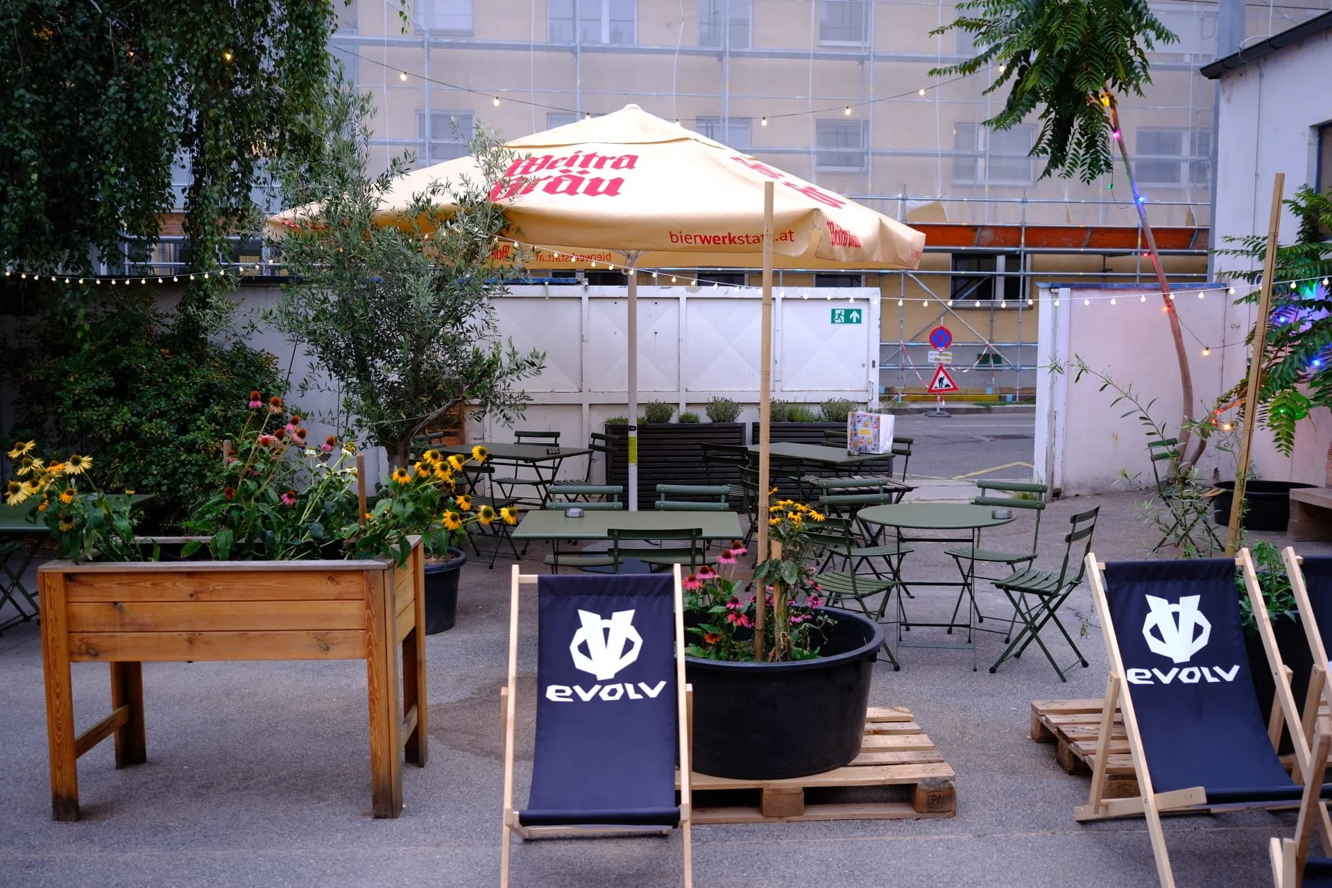 Outdoor patio seating area with tables, chairs, and large planters with flowers, under a large umbrella, surrounded by trees and string lights at dusk.