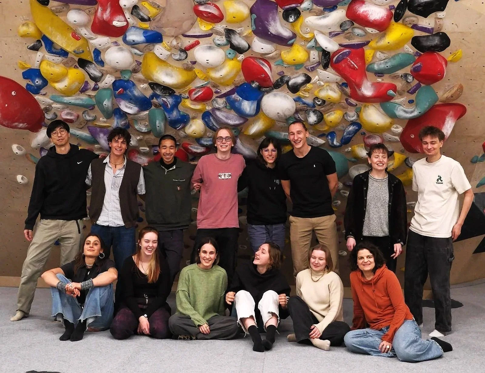 Group of fifteen people posing in front of an indoor climbing wall with colorful holds.