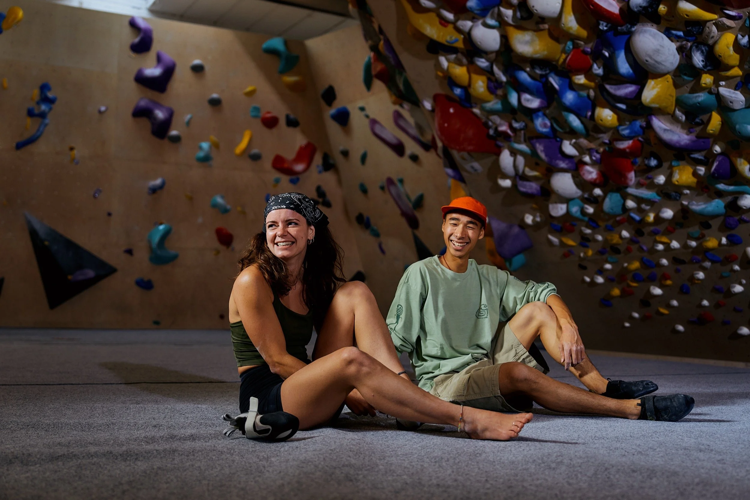 Zwei lachende Menschen, eine Frau und ein Mann, sitzen auf dem Boden in einer Kletterhalle, hinter ihnen bunte Klettergriffe an der Wand.
